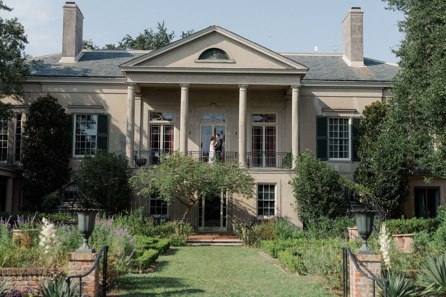 Elegant beige colonial style mansion with multiple wings and chimneys set among landscaped gardens and mature trees.