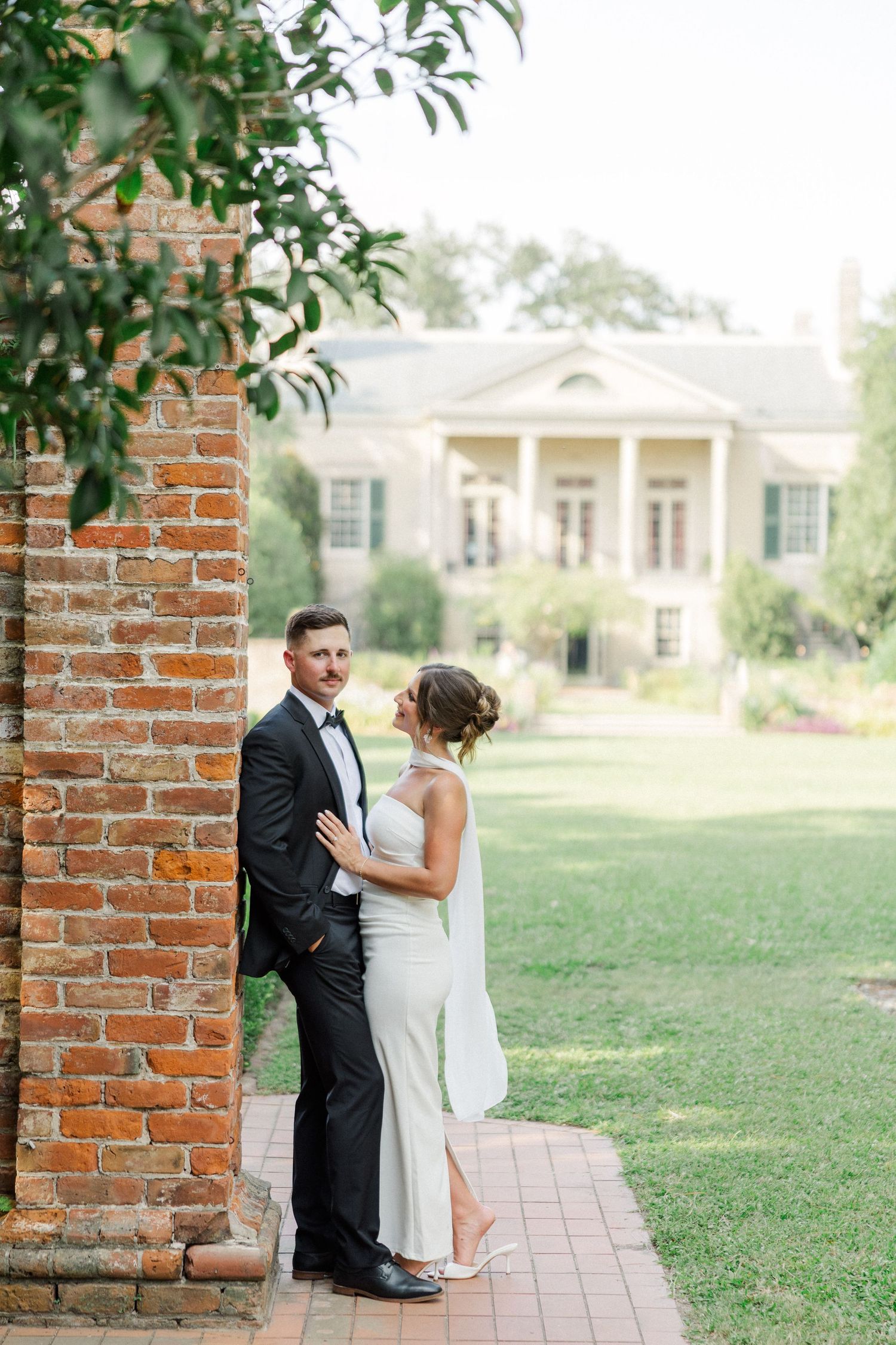 A bride and groom share an intimate moment by a brick column with a stately mansion visible in the background.