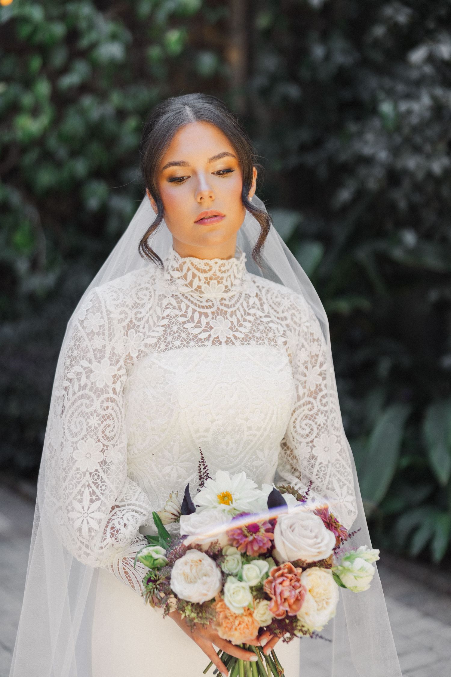 A bride in a high-neck lace wedding gown holds an elegant bouquet of mixed roses and flowers against a natural backdrop.