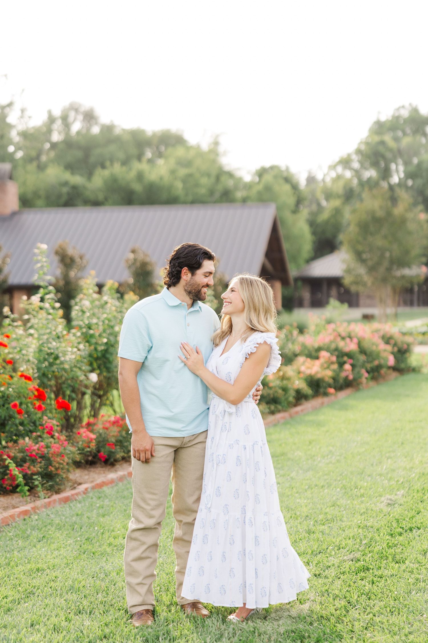 A couple shares a tender moment while standing in a beautiful garden with red flowers and rustic buildings.