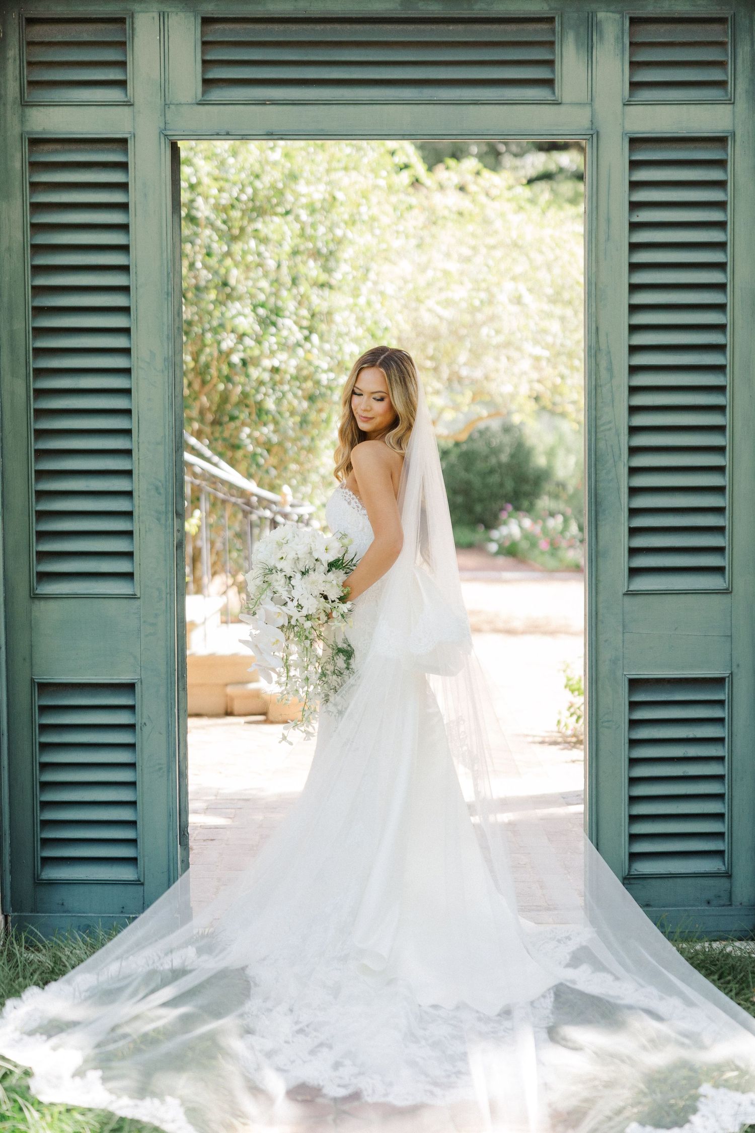 Romantic bridal portrait framed by vintage green shutters showing flowing cathedral length veil.
