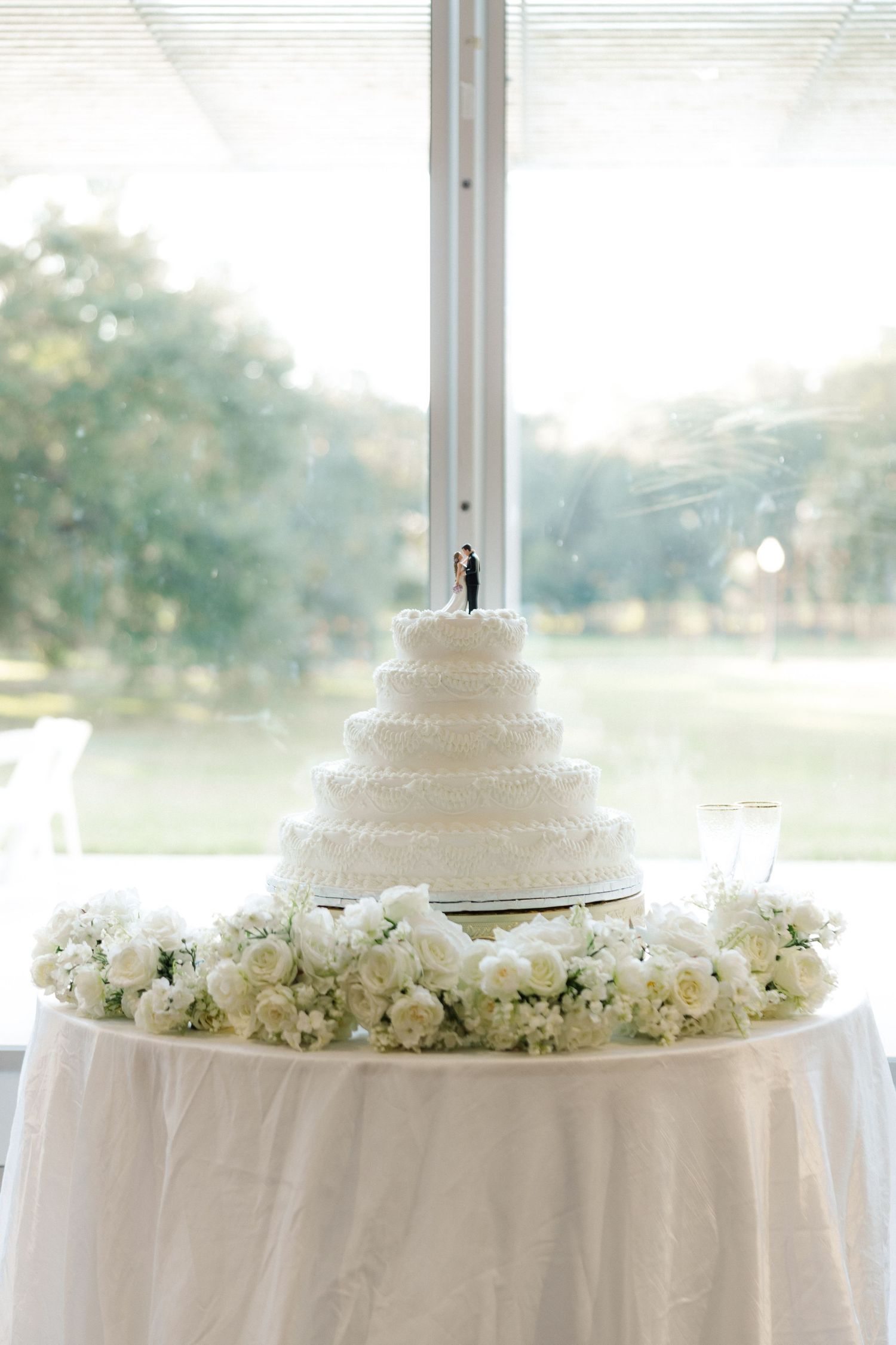 White tiered wedding cake displayed on table with floral arrangements and white tablecloth.