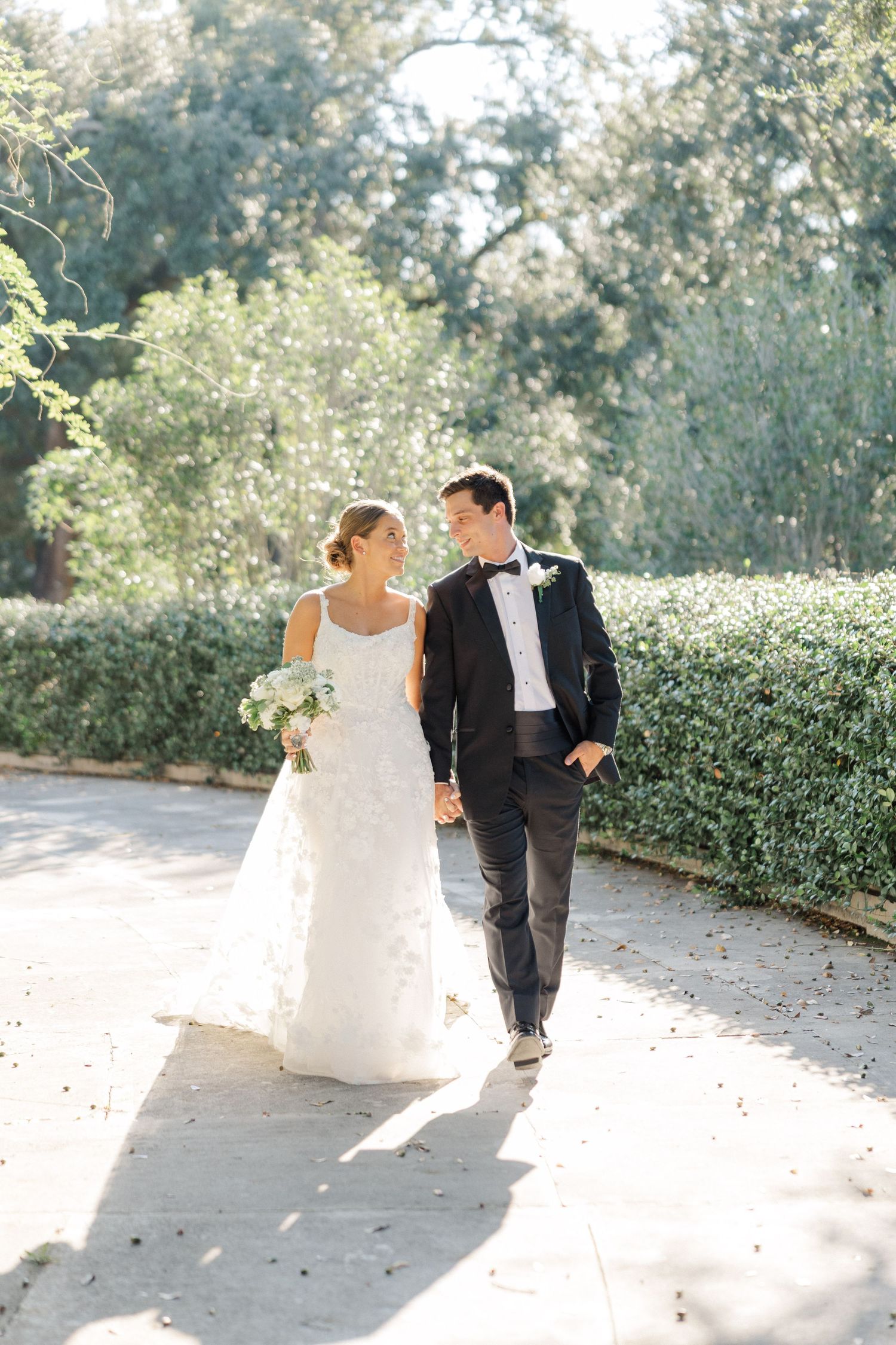 Newlyweds walking together on a sunlit garden path with flowing white dress and classic tuxedo.