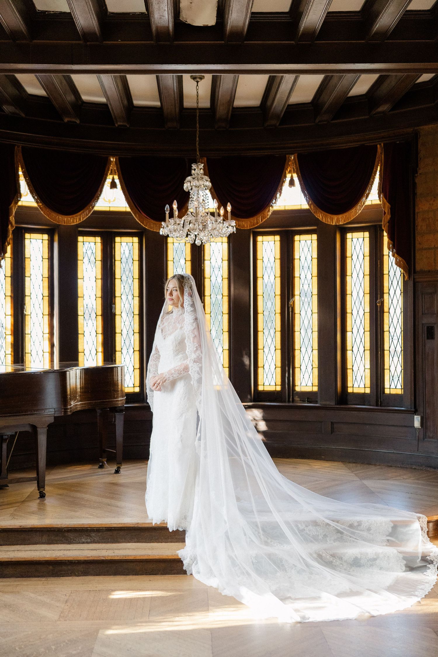 Long flowing wedding veil trails across wooden floor in sunlit room with stained glass windows.