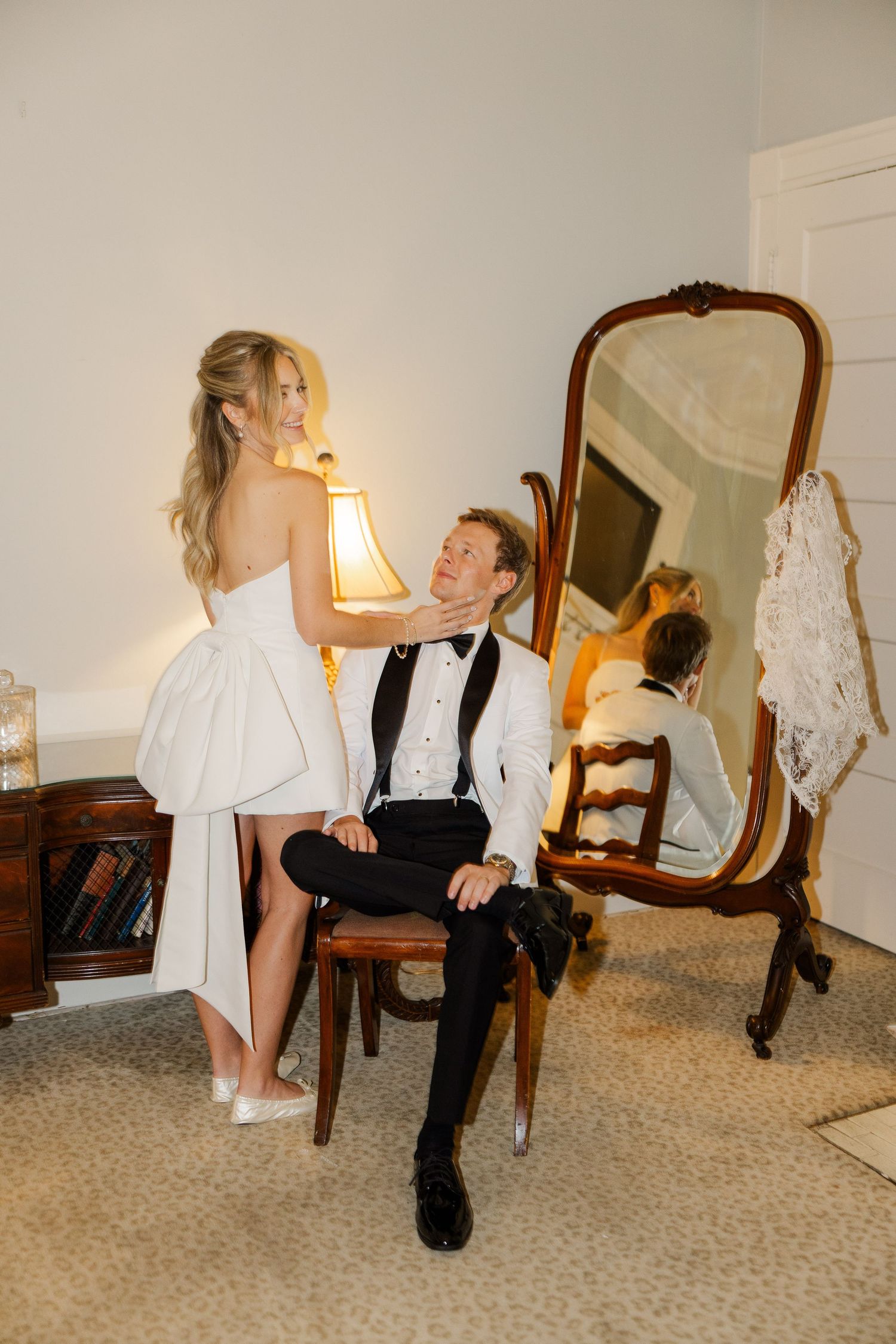 A couple poses playfully in a bedroom setting with antique furniture and soft lighting for wedding photos.