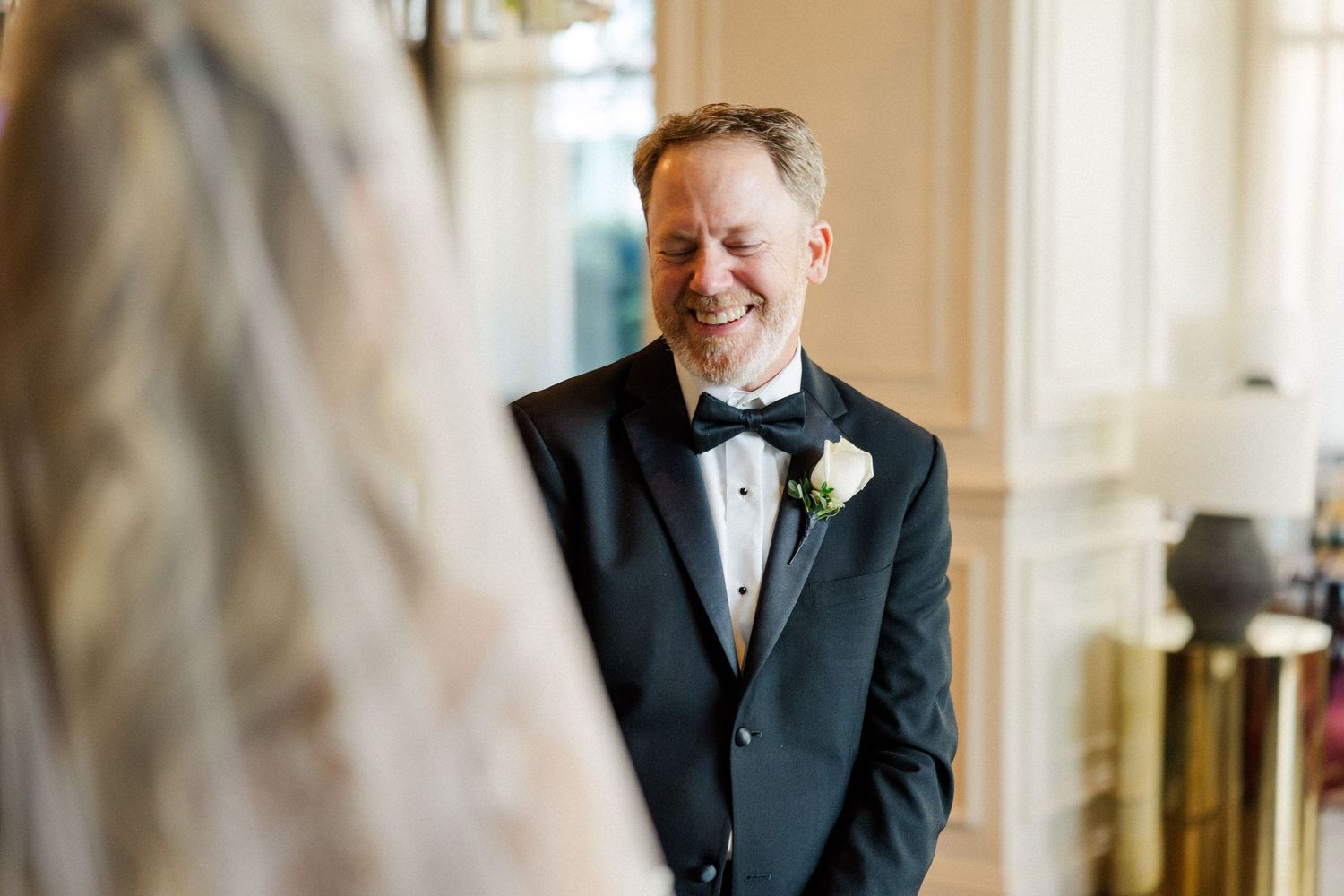 A groom in a classic black tuxedo and bow tie smiles while getting ready in an elegant room with natural lighting.