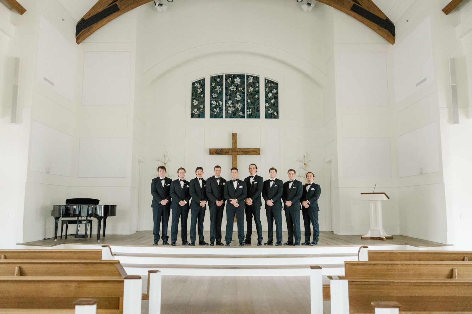 Wedding party members stand in formal attire inside bright white church sanctuary.