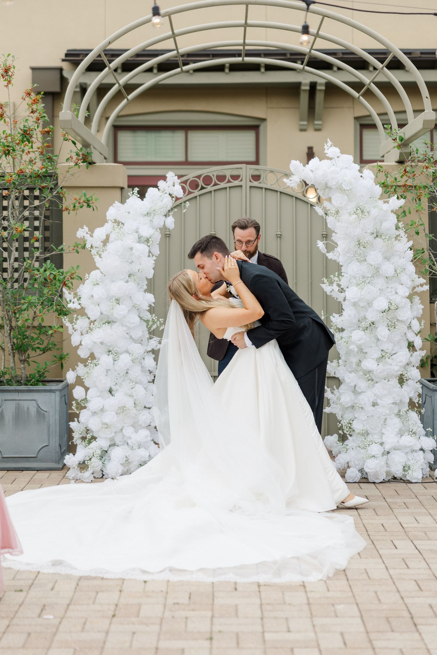 Wedding couple shares romantic moment under white floral arch in courtyard setting.