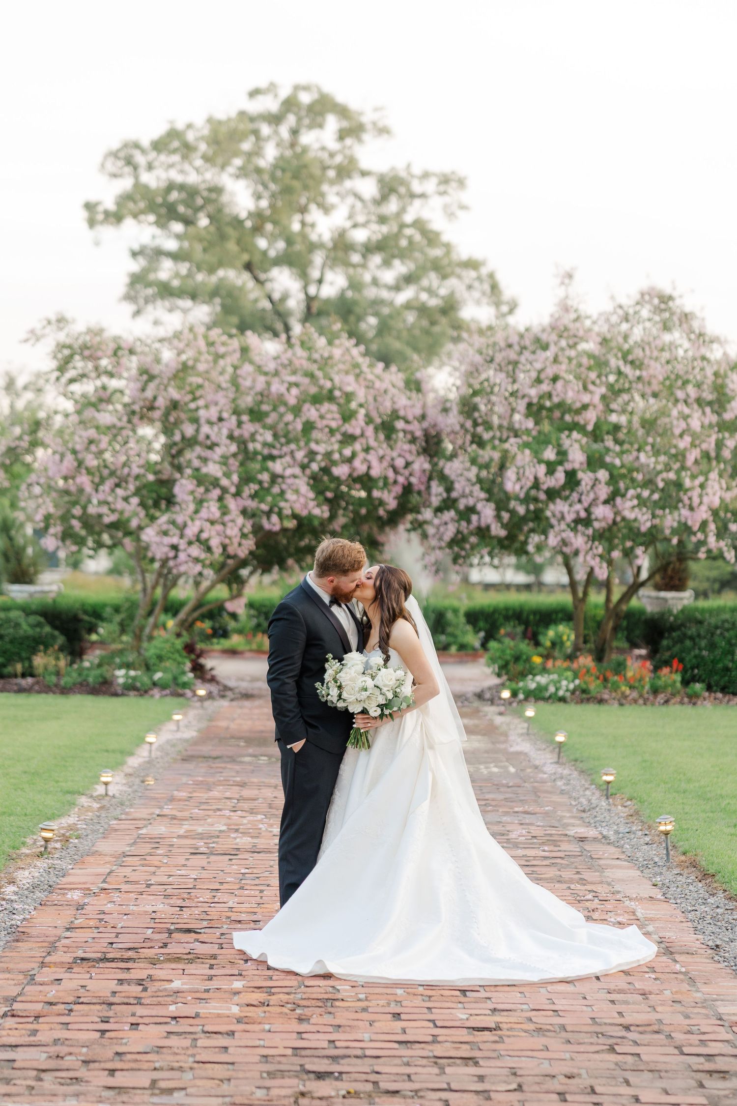 A couple shares an intimate moment on a brick pathway surrounded by blooming cherry blossom trees in spring.