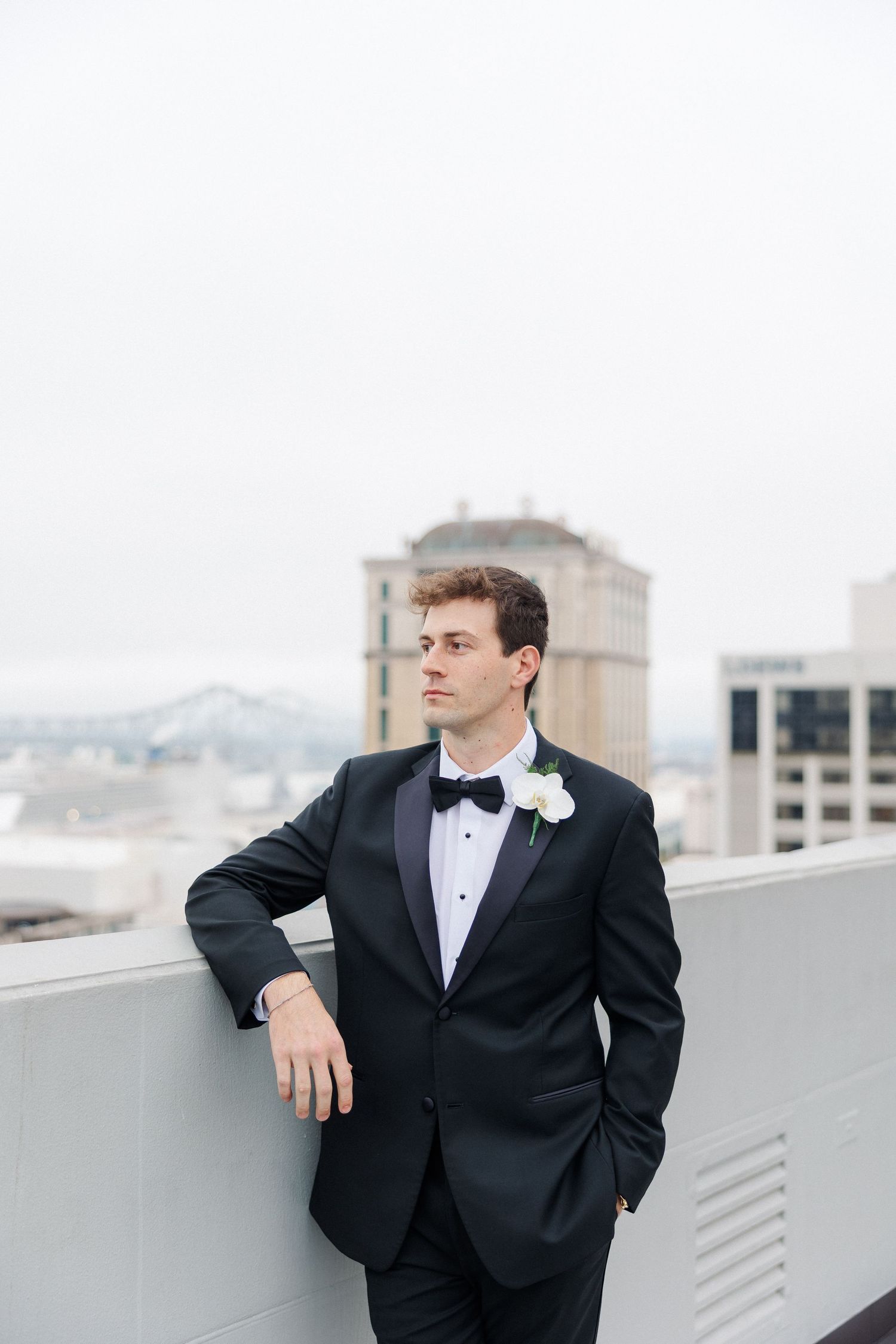 Elegant groom in black tuxedo poses on rooftop with city skyline backdrop.