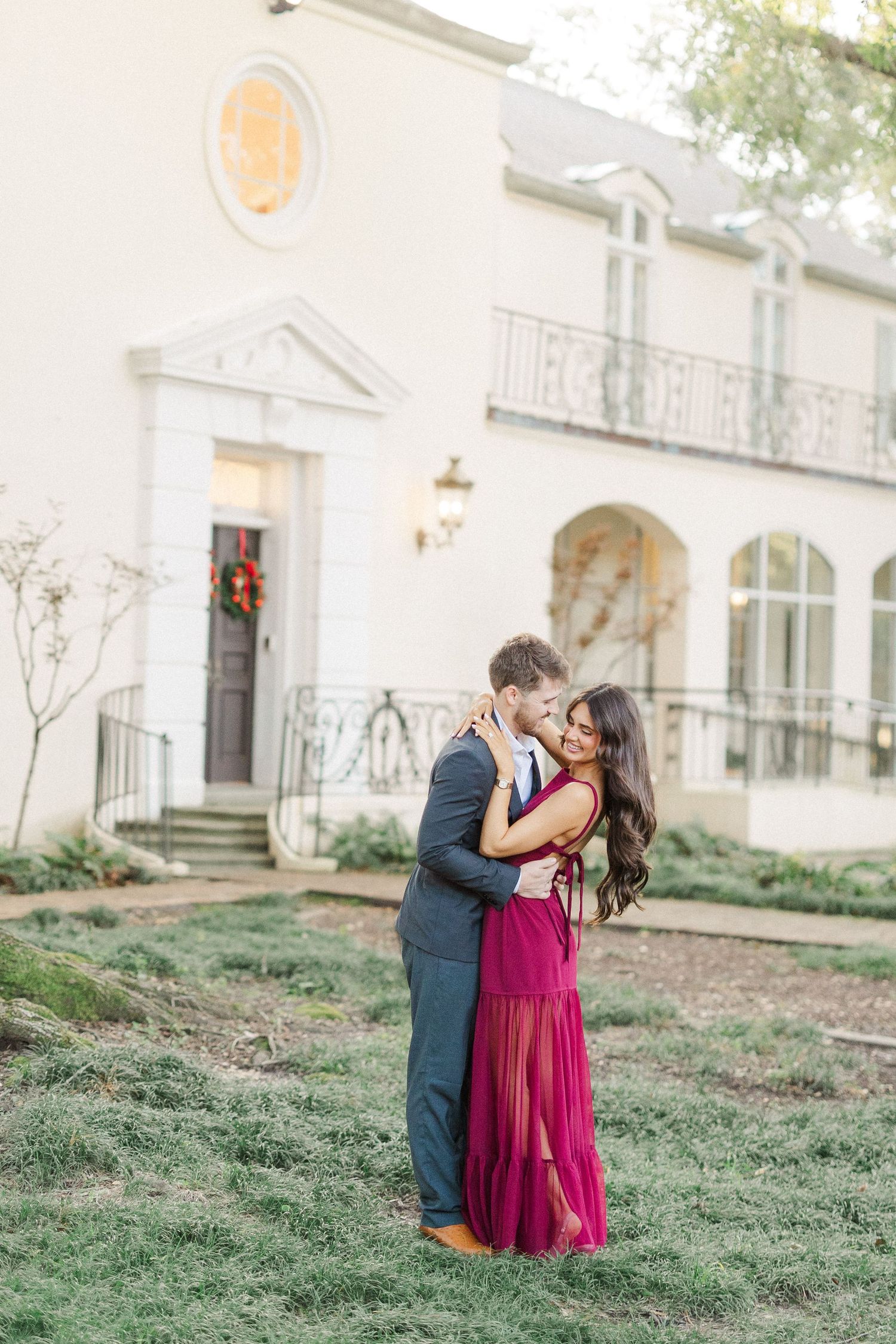 A couple shares a romantic moment on a grassy slope in front of a white colonial-style building at sunset.