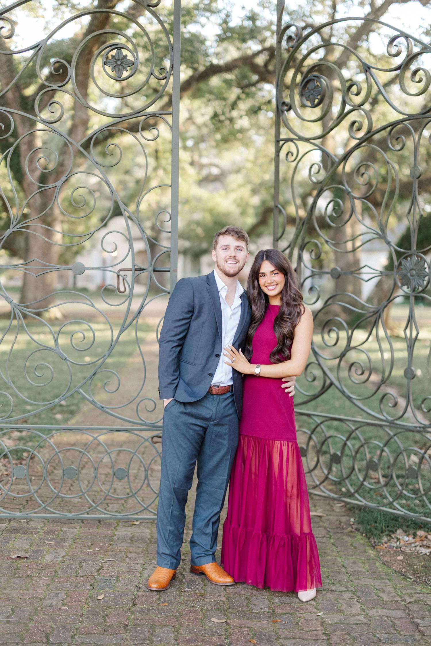 A couple poses together against a decorative iron gate in an outdoor setting, with her wearing a burgundy dress.