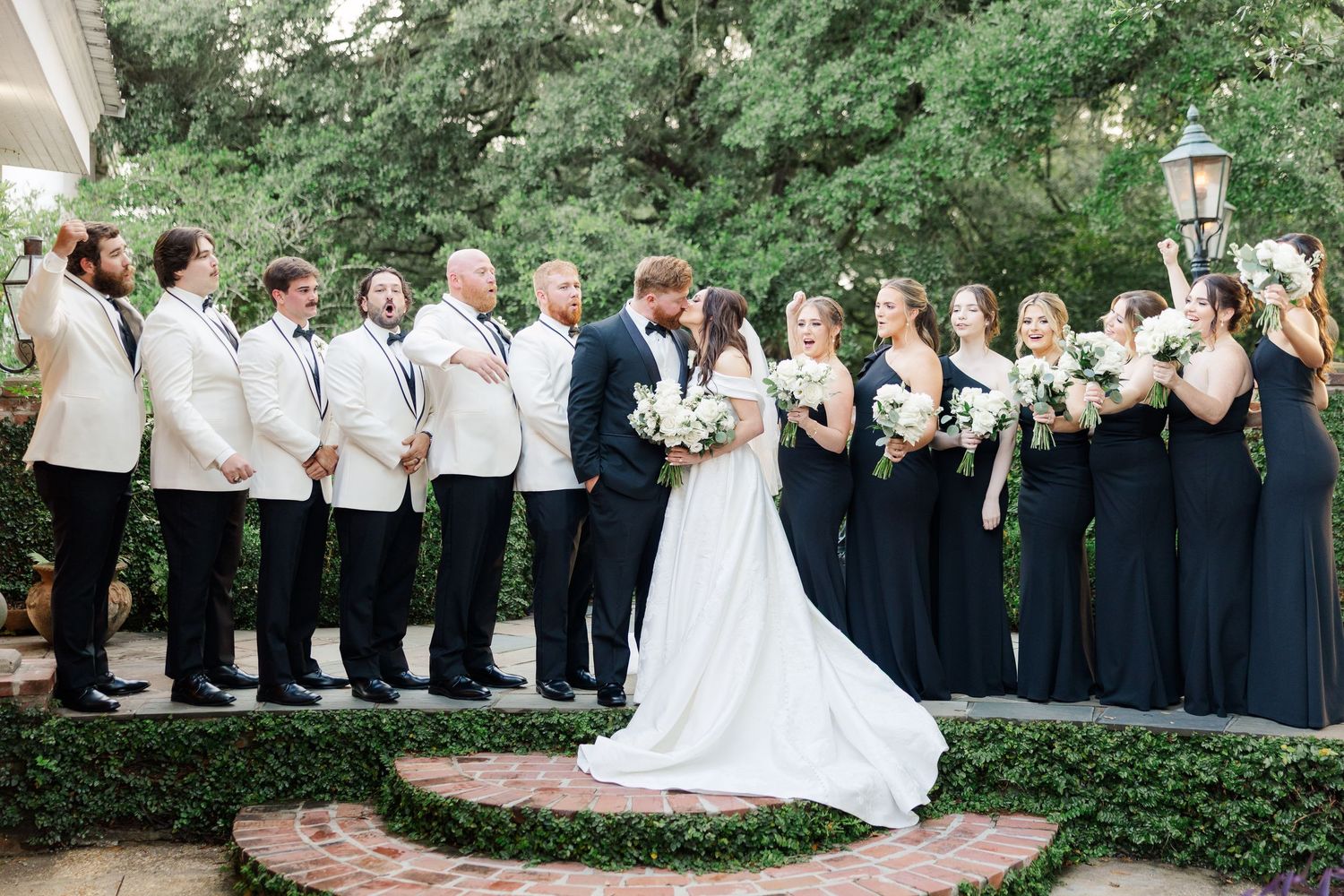 Wedding party in white and navy blue attire poses outdoors in a garden setting with trees and greenery.