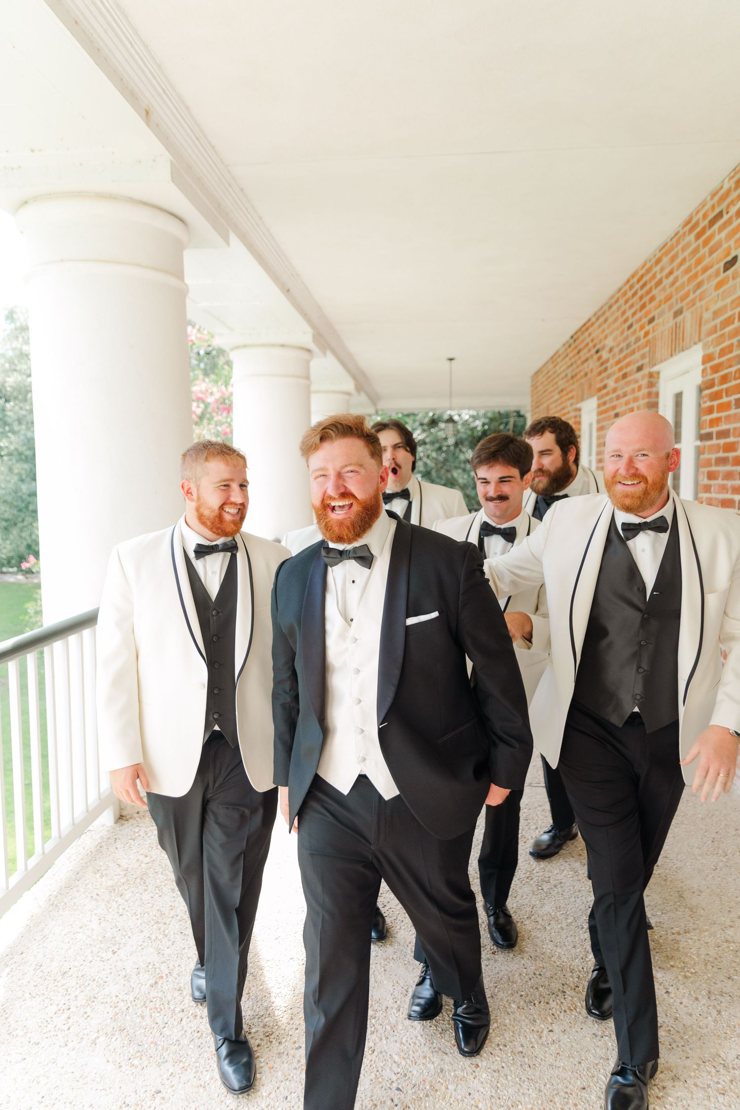 Groomsmen in black tuxedos and white vests walk along a columned brick porch.