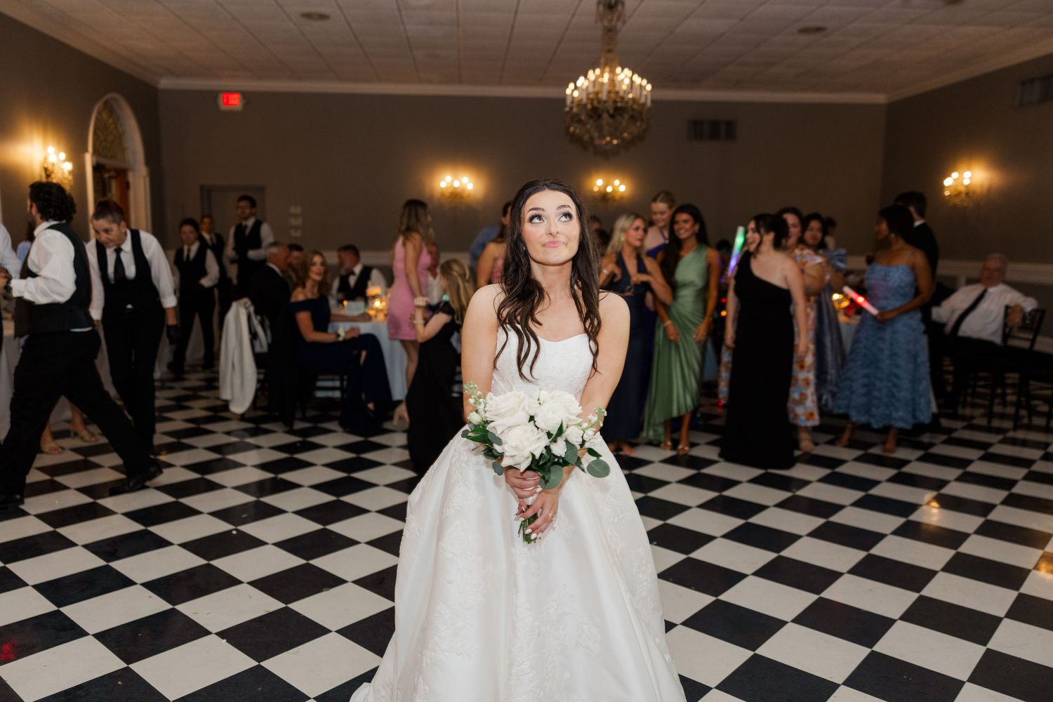Wedding reception guests dance on a black and white checkered dance floor under warm lighting.