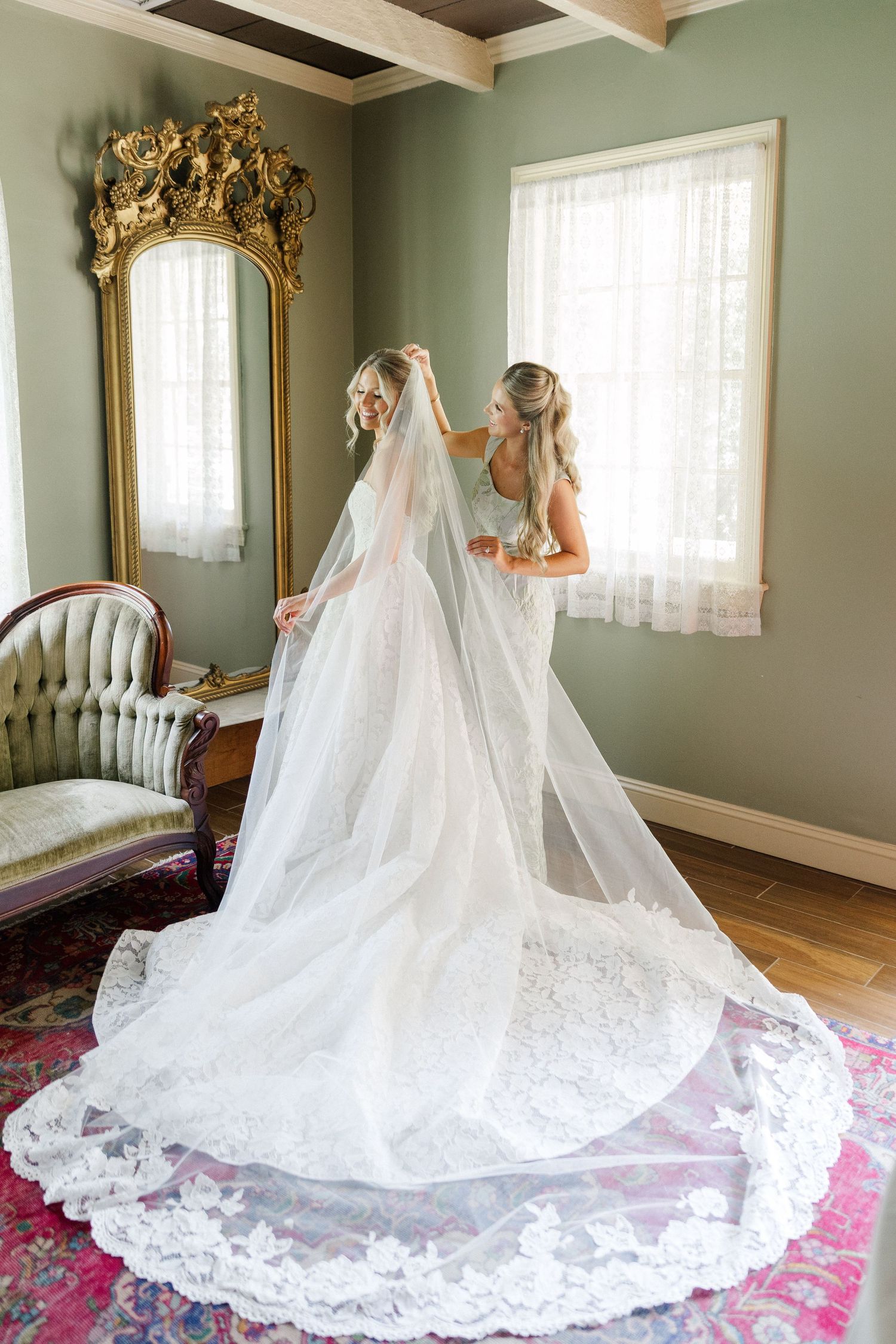 Bride in lace gown with cathedral veil being adjusted in a vintage green room with gilded mirror.