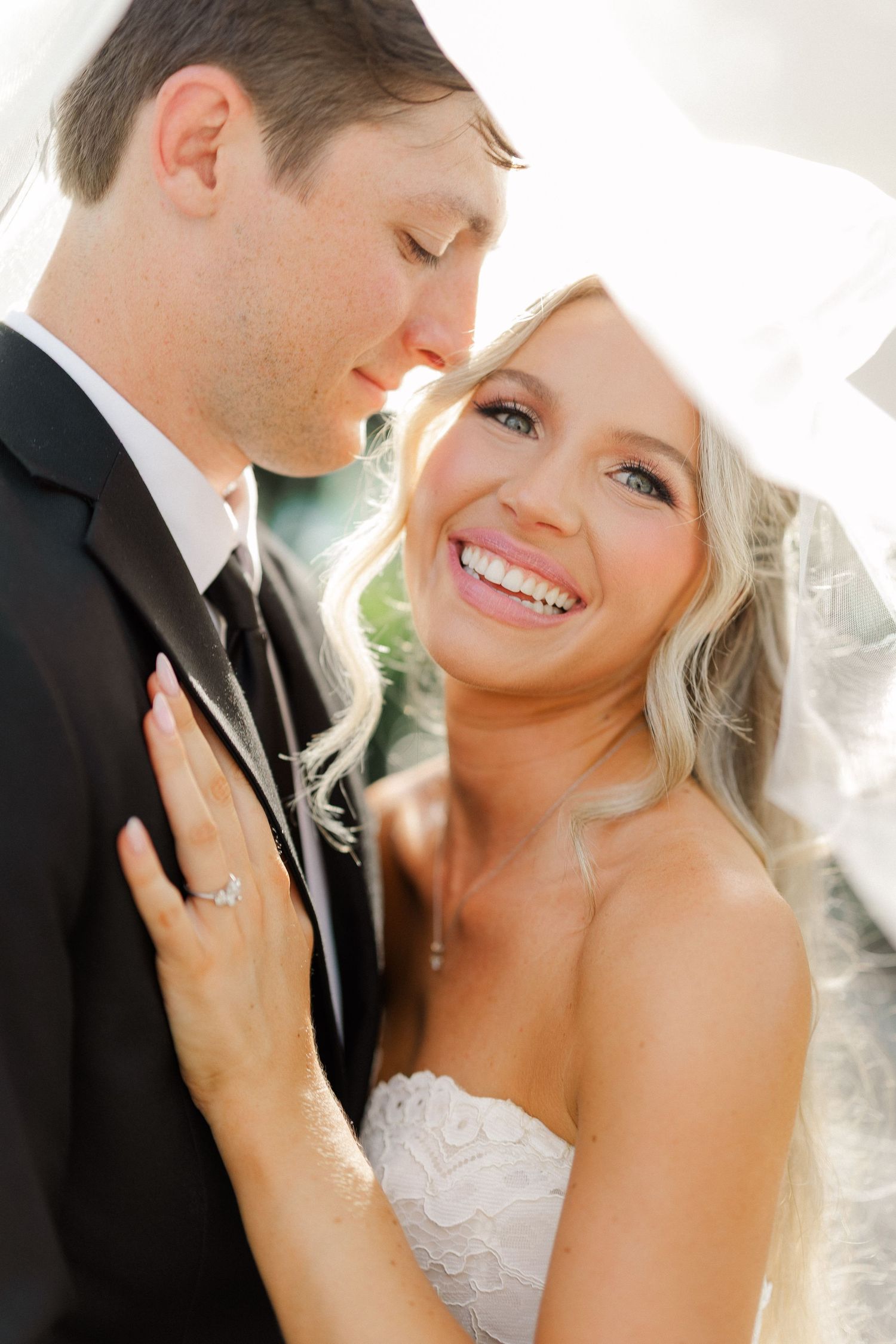 Smiling bride in strapless lace dress leans close to groom under veil in romantic wedding portrait.