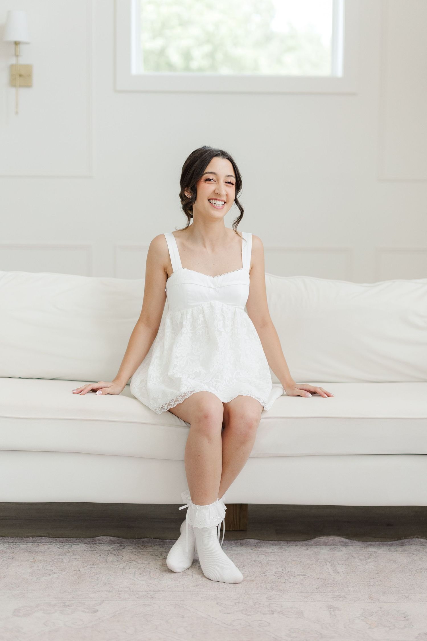 Smiling bride in white mini dress and white boots sitting on a cream sofa in a bright white room.