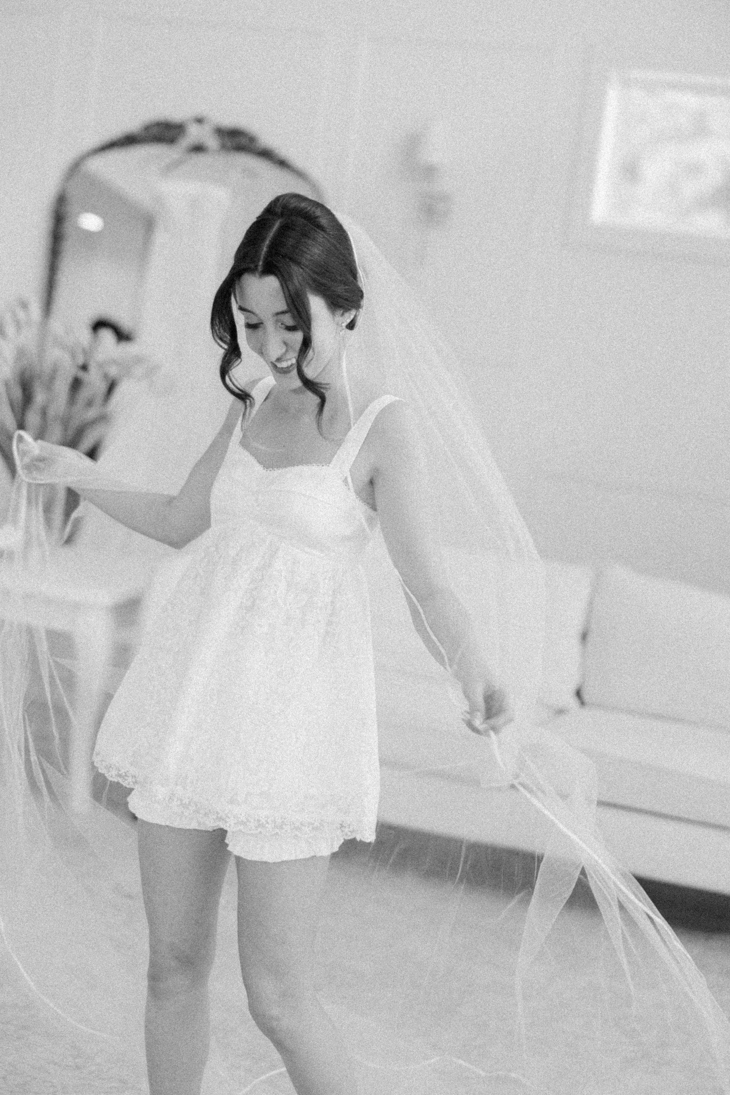 Black and white photo of bride in white mini dress holding flowing veil in bridal suite.