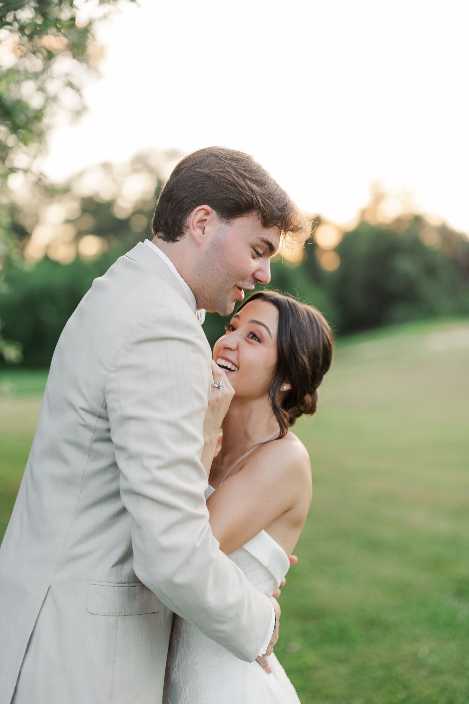 Groom in tan suit tenderly embraces smiling bride in white strapless gown at golden hour outdoors.