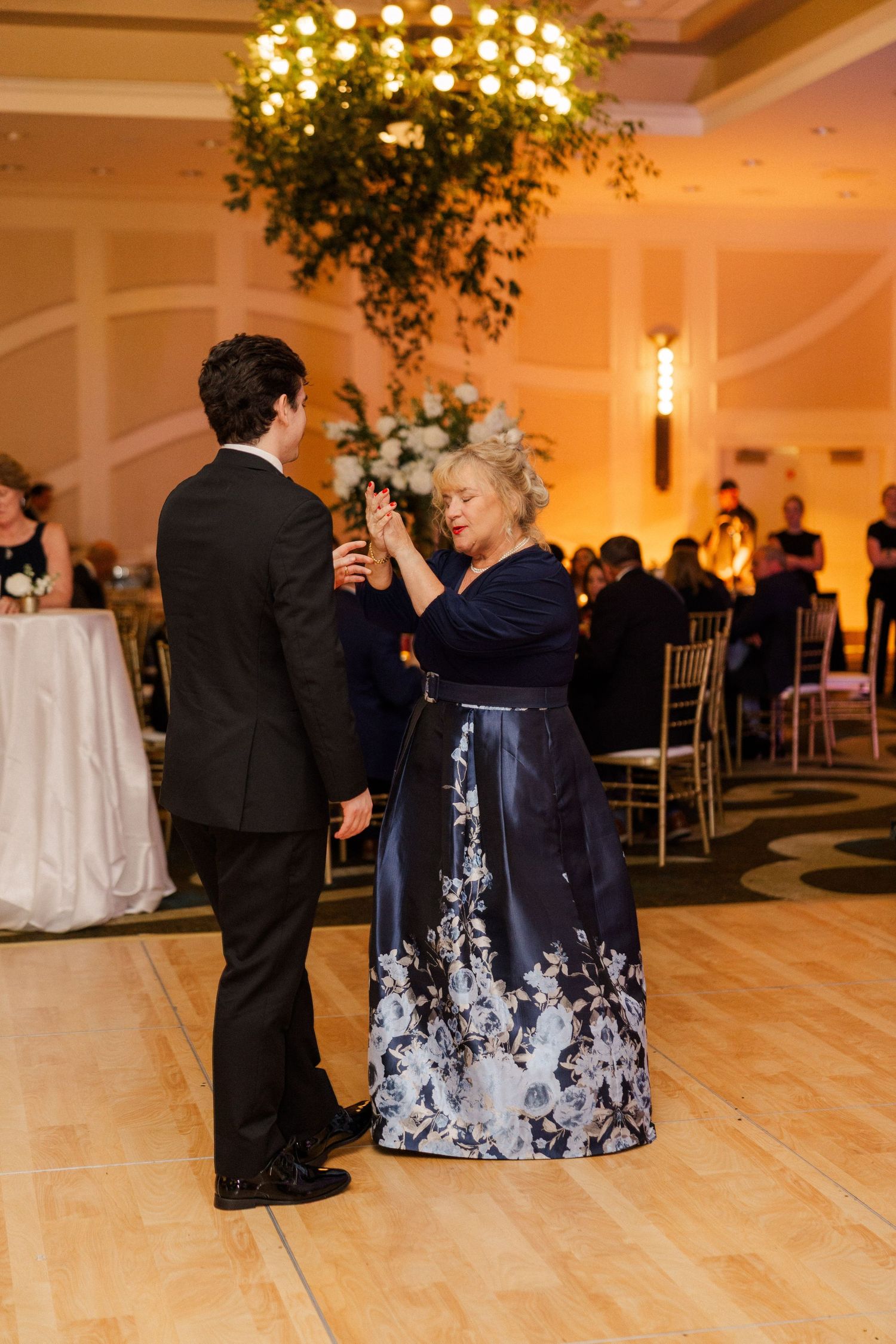 A groom dances with his mother in a navy floral gown at an elegant wedding reception.