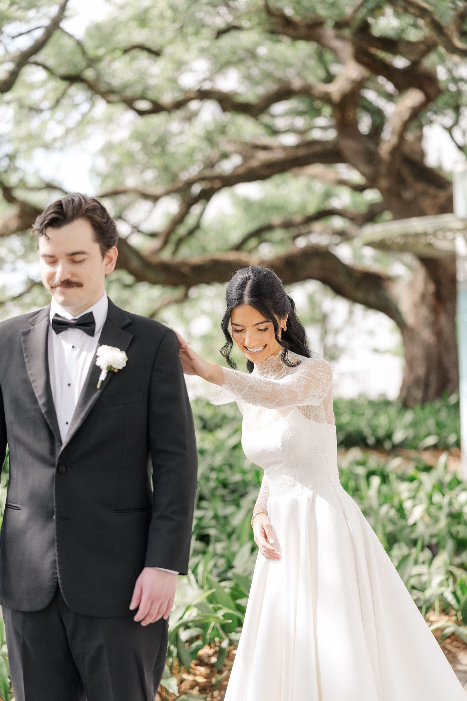 Bride in lace gown touches grooms shoulder during outdoor first look under oak tree.