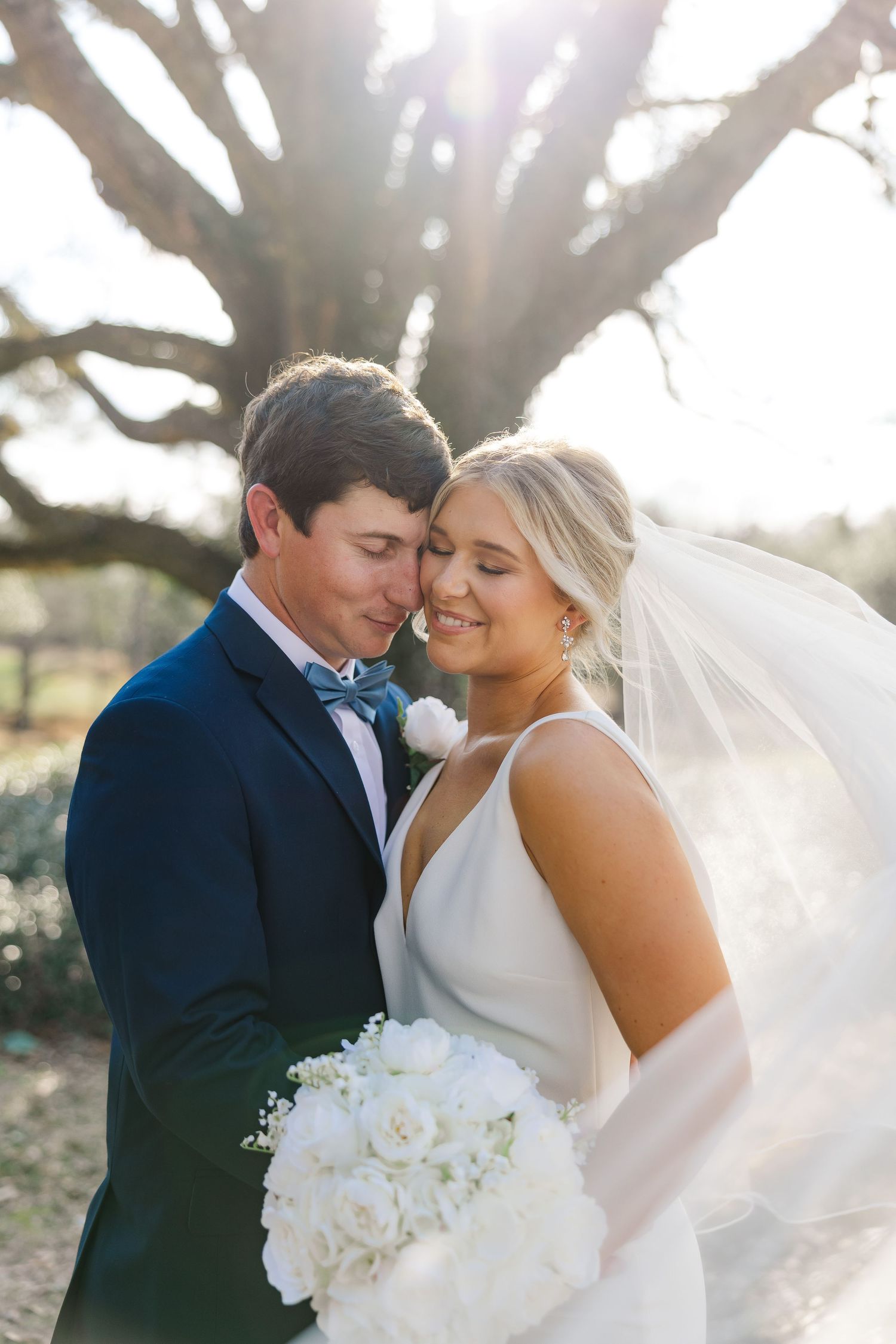 A romantic outdoor wedding moment with sunlight filtering through trees as couple smiles with white bouquet.