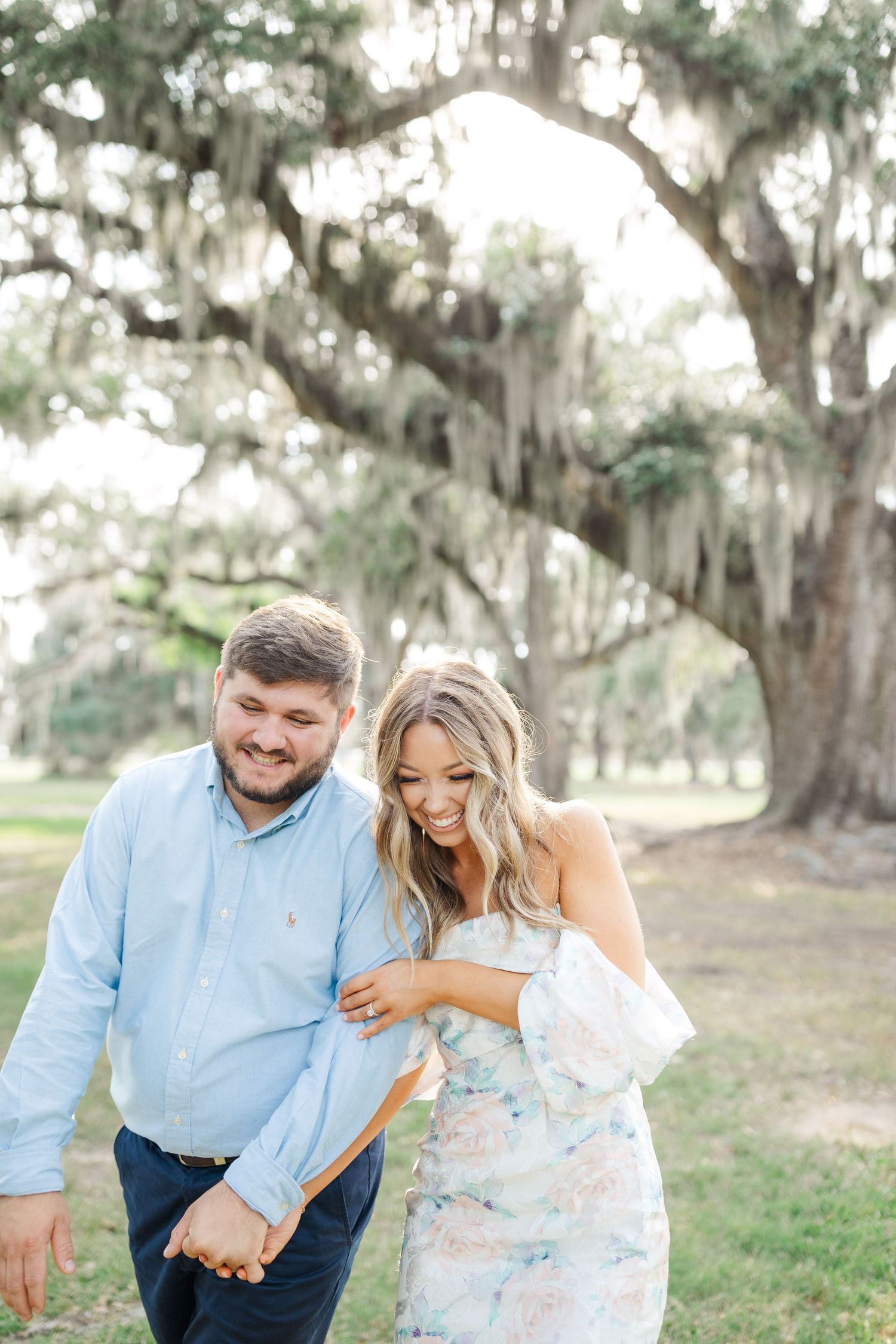A couple shares a joyful laugh while walking together under Spanish moss-draped oak trees in a romantic outdoor setting.
