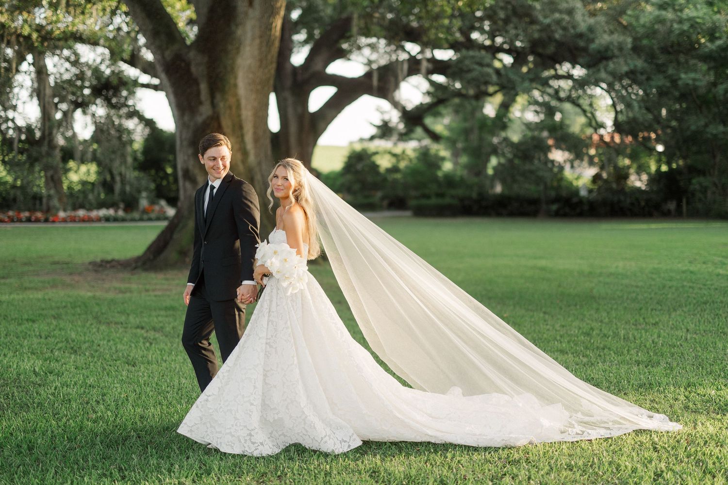 Bride in flowing white gown with long veil walks with groom in black tux on lush green lawn near large oak tree.