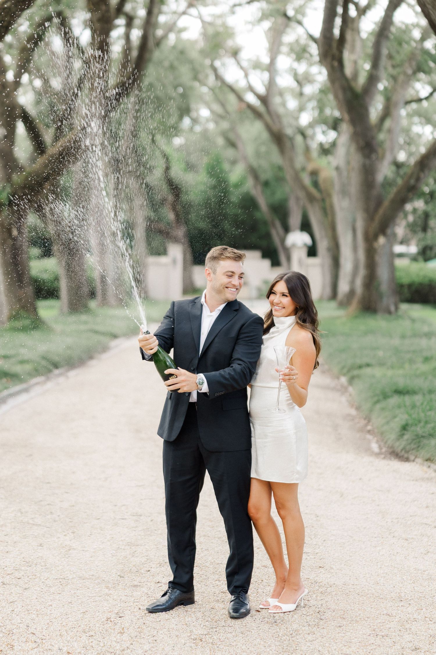 Couple celebrating engagement by popping champagne on a tree-lined path, woman in white dress, man in black suit.