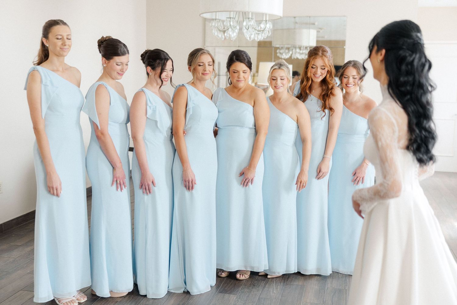Bride in white gown faces seven bridesmaids in light blue floor-length dresses during a bright indoor first look moment.