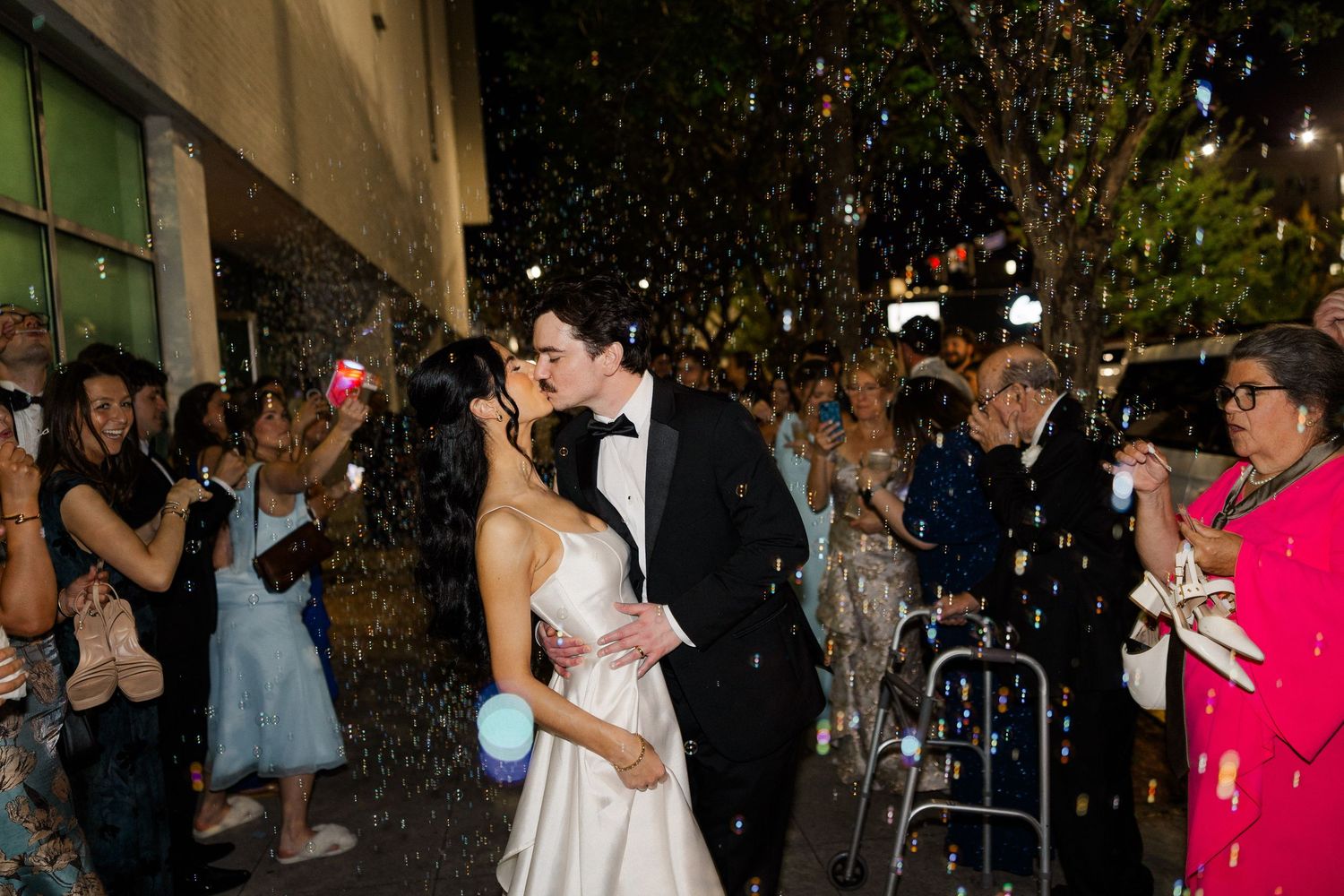Bride and groom share a kiss during confetti send-off after outdoor wedding ceremony.