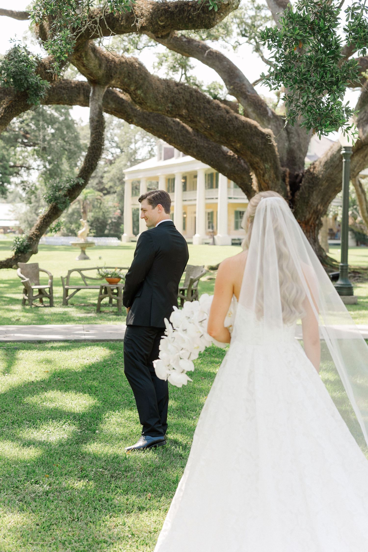Bride in white gown with veil and groom in black tux share first look under sprawling oak tree at elegant estate.