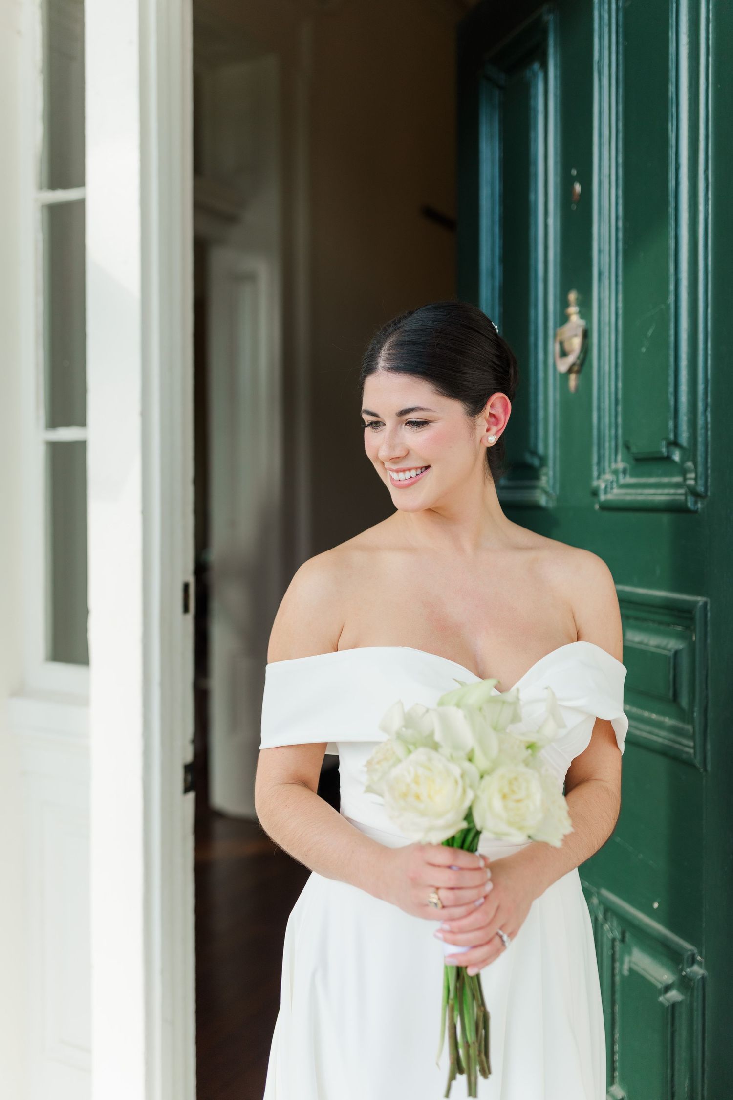 Elegant bride in off-shoulder white dress smiles while holding white roses beside a green vintage door.