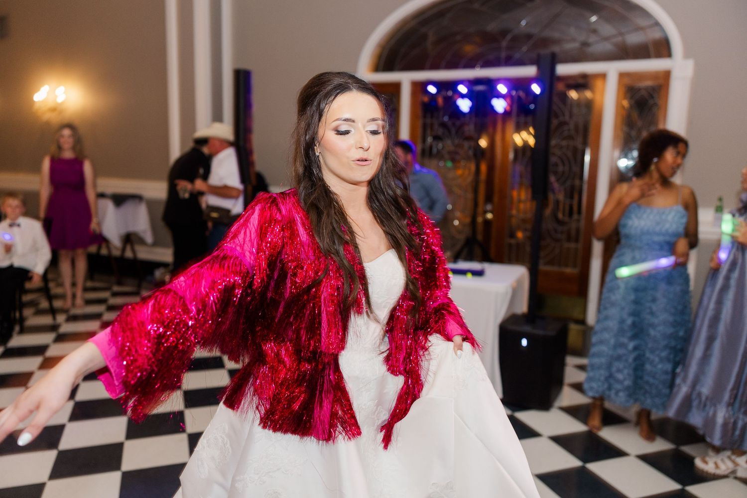 A bride wearing a sparkling red jacket twirls on a black and white checkered dance floor at her wedding reception.