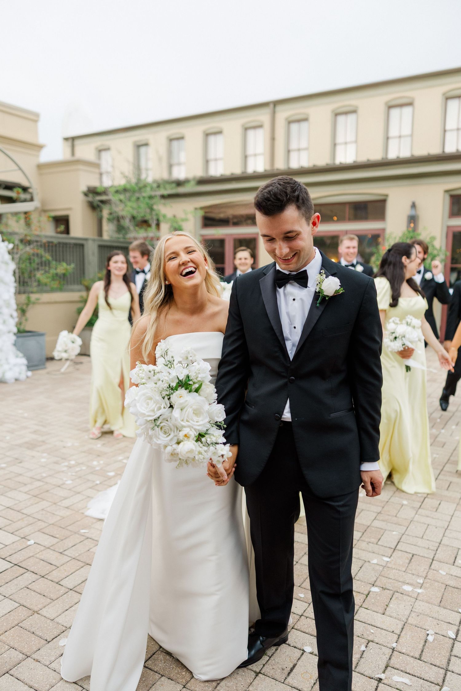 Joyful wedding couple shares candid laughs while walking together on a stone patio in formal attire.
