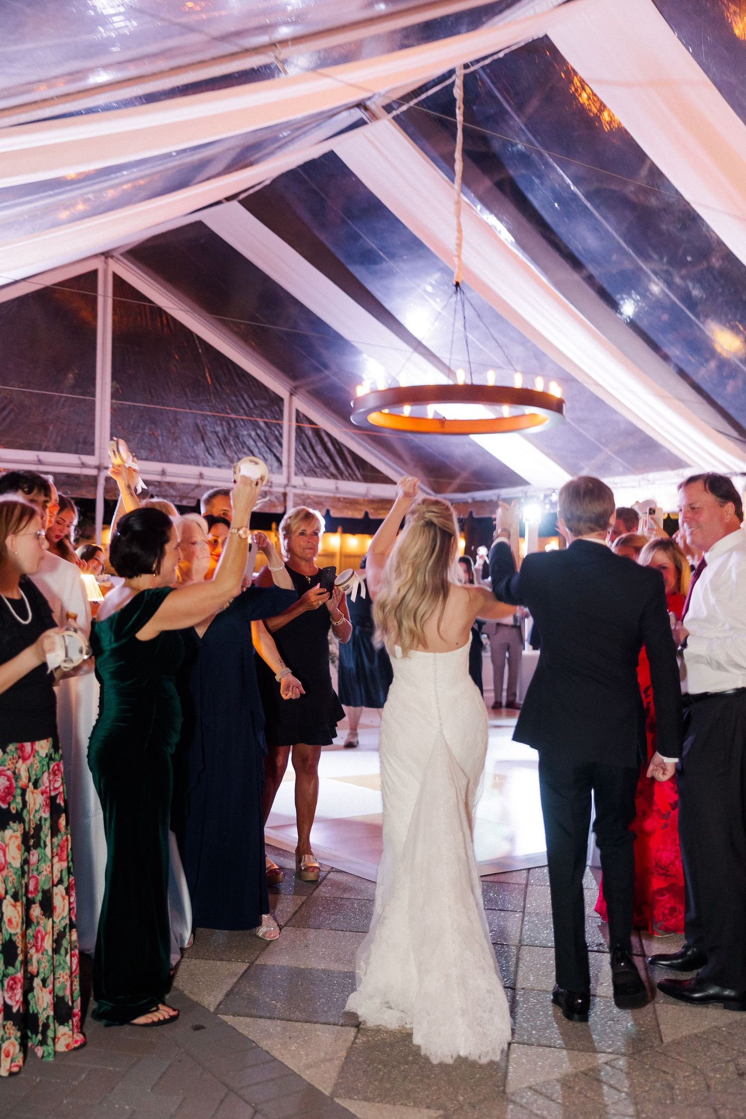 Wedding guests and couple dancing together under a white tent with festive lighting.
