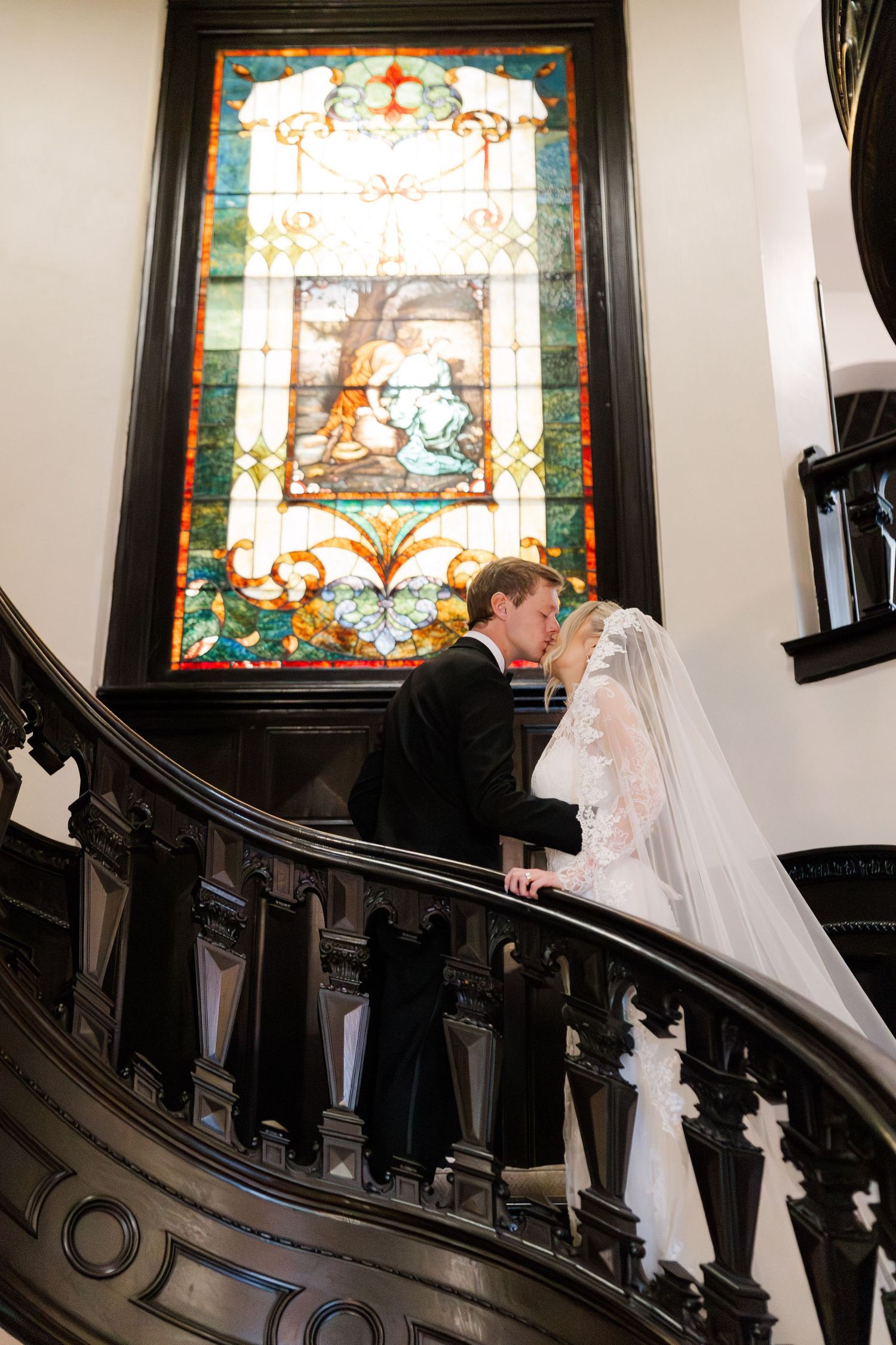 Wedding couple ascending elegant staircase with stained glass windows in historic venue.