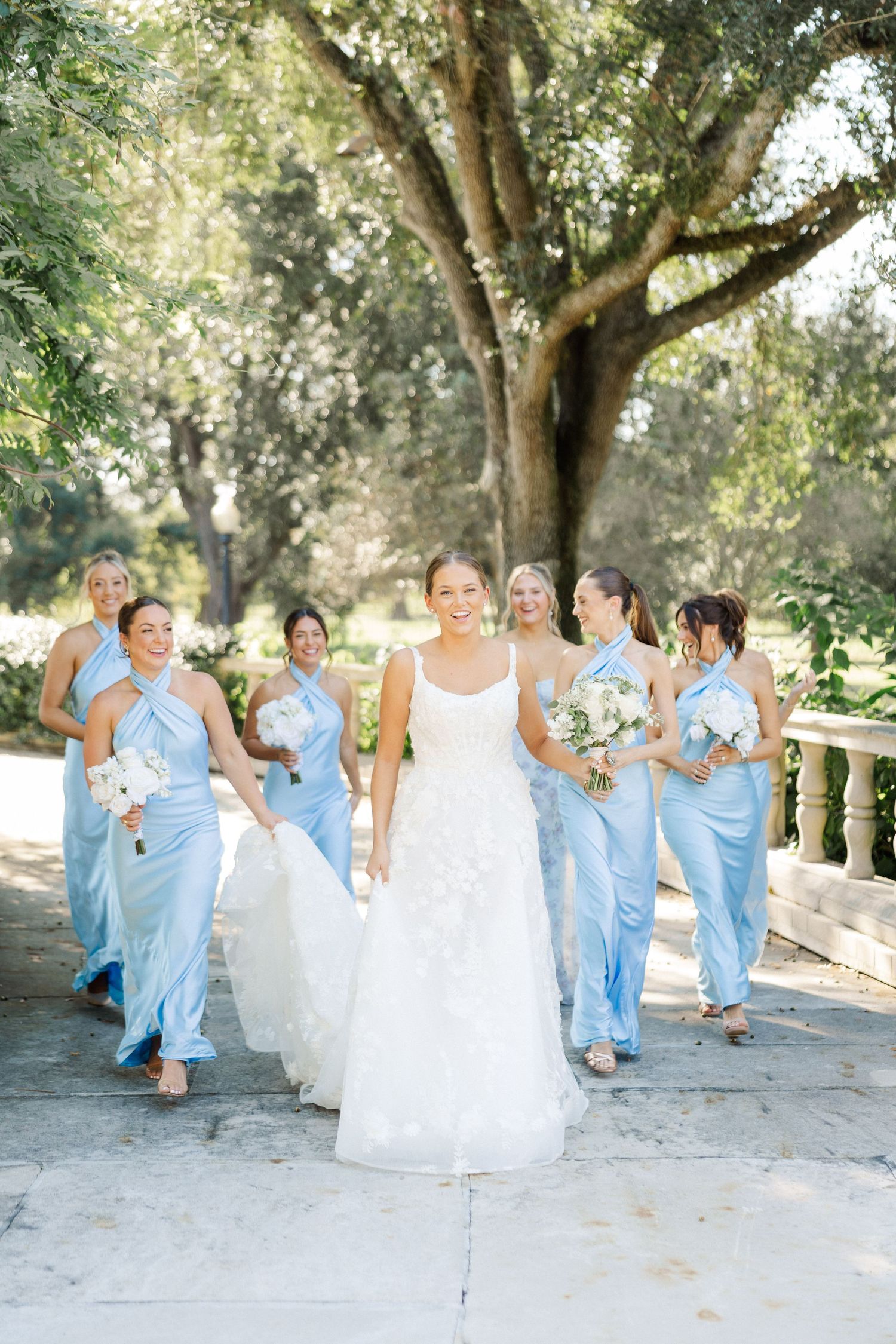 Bridal party in light blue dresses runs playfully along a stone pathway outdoors.