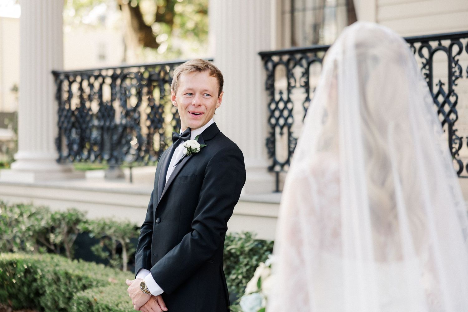 A groom in a black tuxedo smiles during a first look moment with his bride.