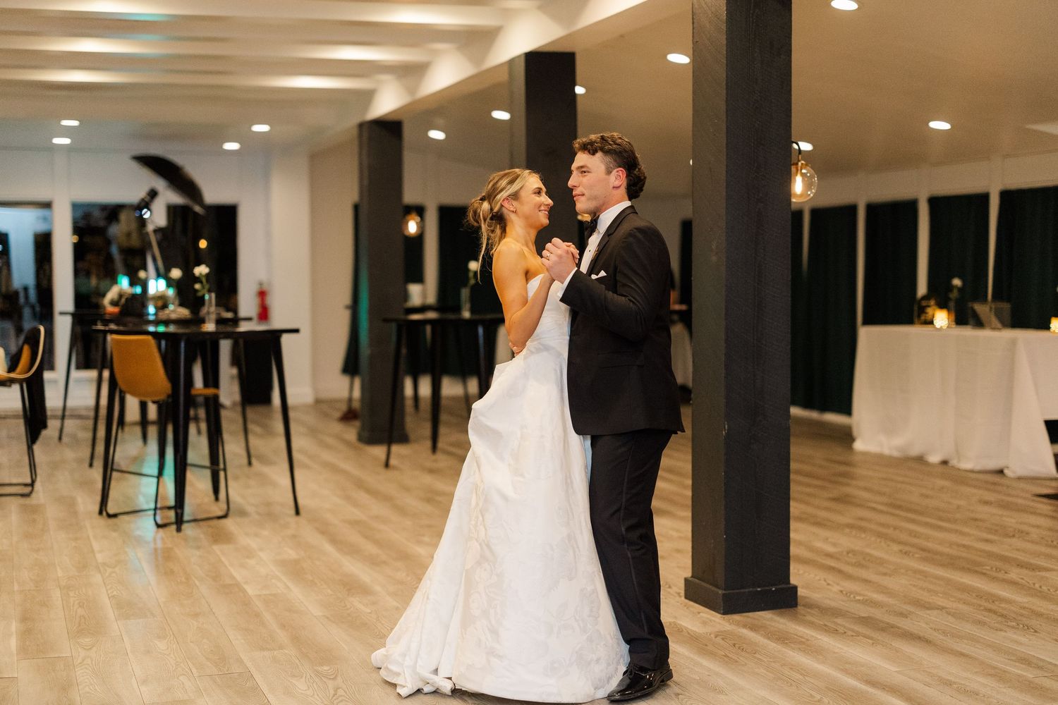 Wedding couple dancing in modern reception venue with dark pillars and light wood floors.