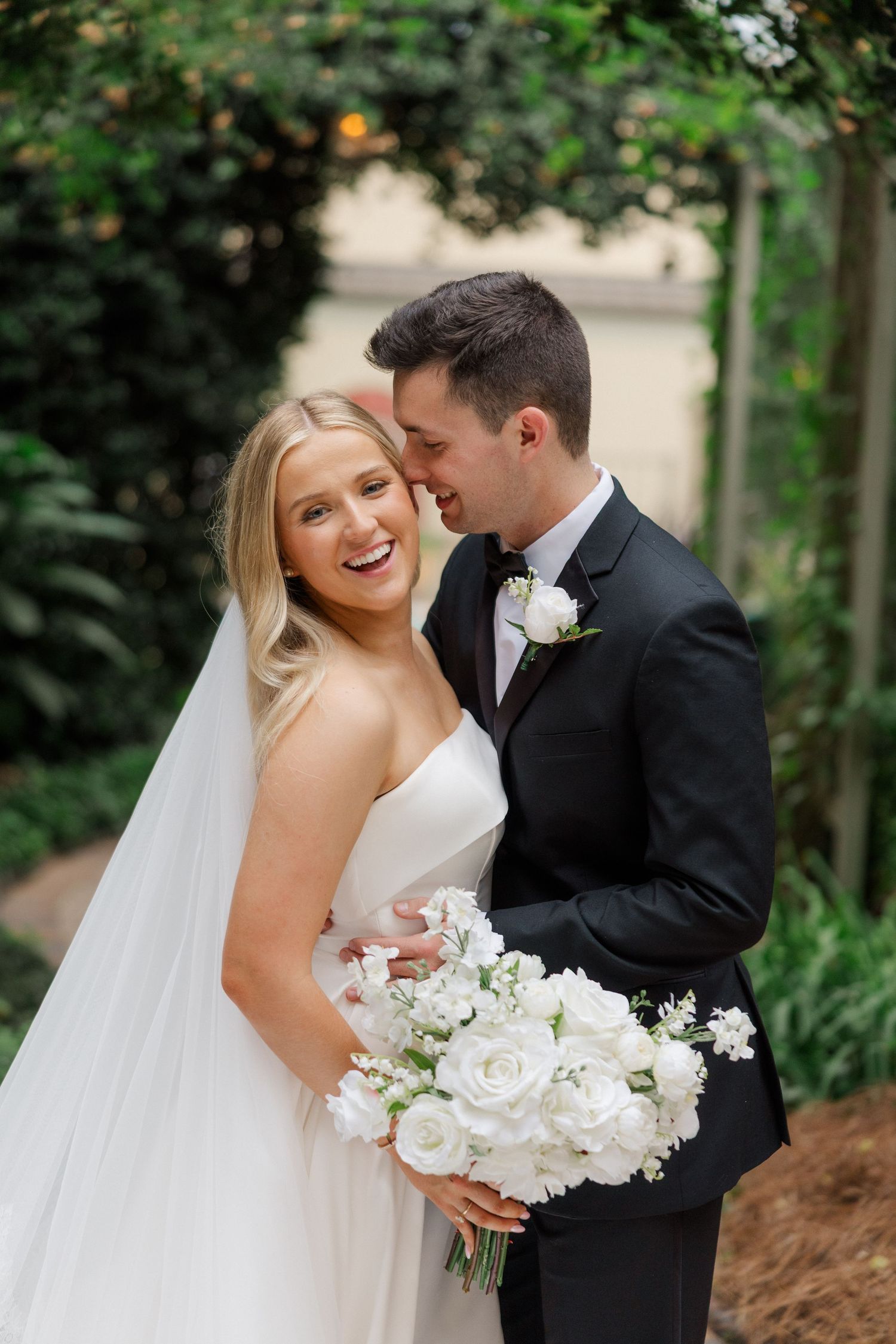 Newlyweds laughing together in a garden setting with white floral bouquet.