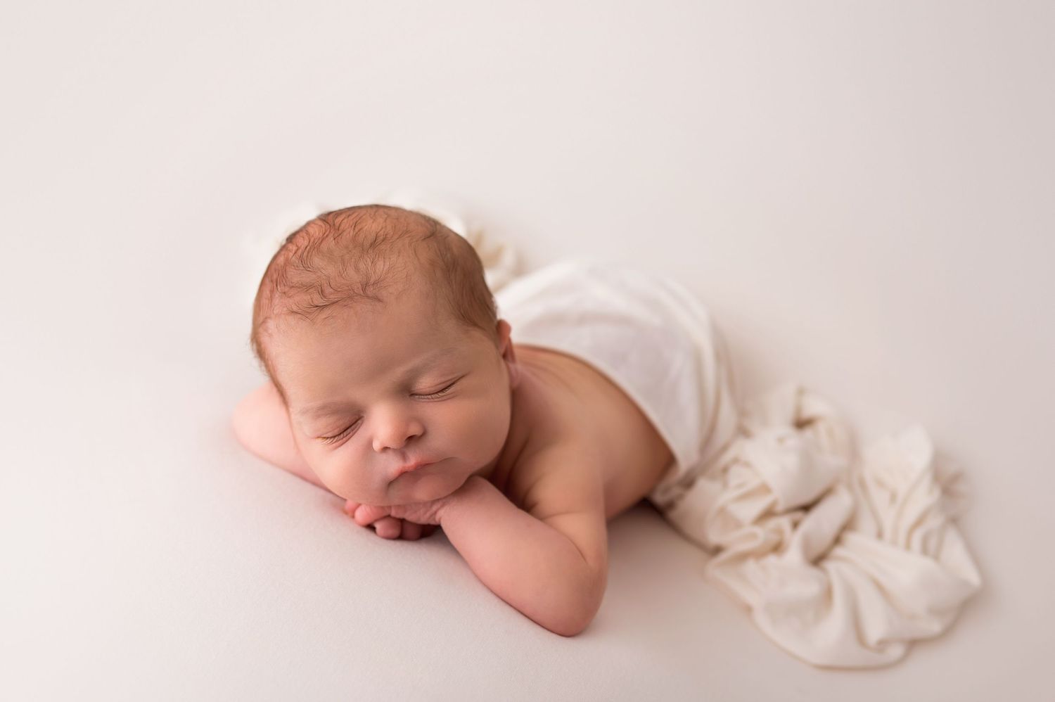 Peaceful sleeping newborn baby lying on white backdrop with soft white blanket during professional studio photography session.