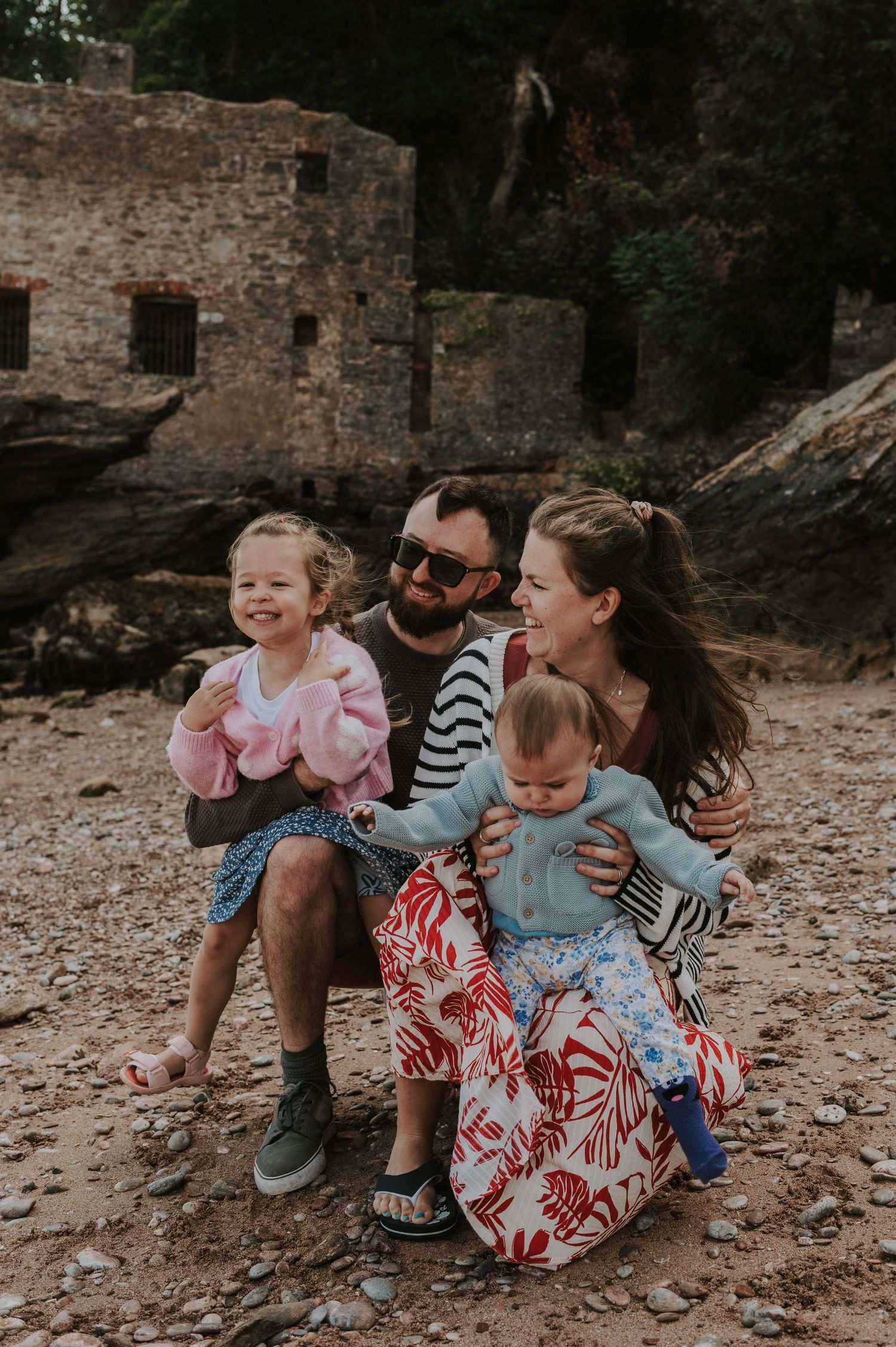 Family enjoys a candid moment together on a rocky beach near old stone ruins during sunset.