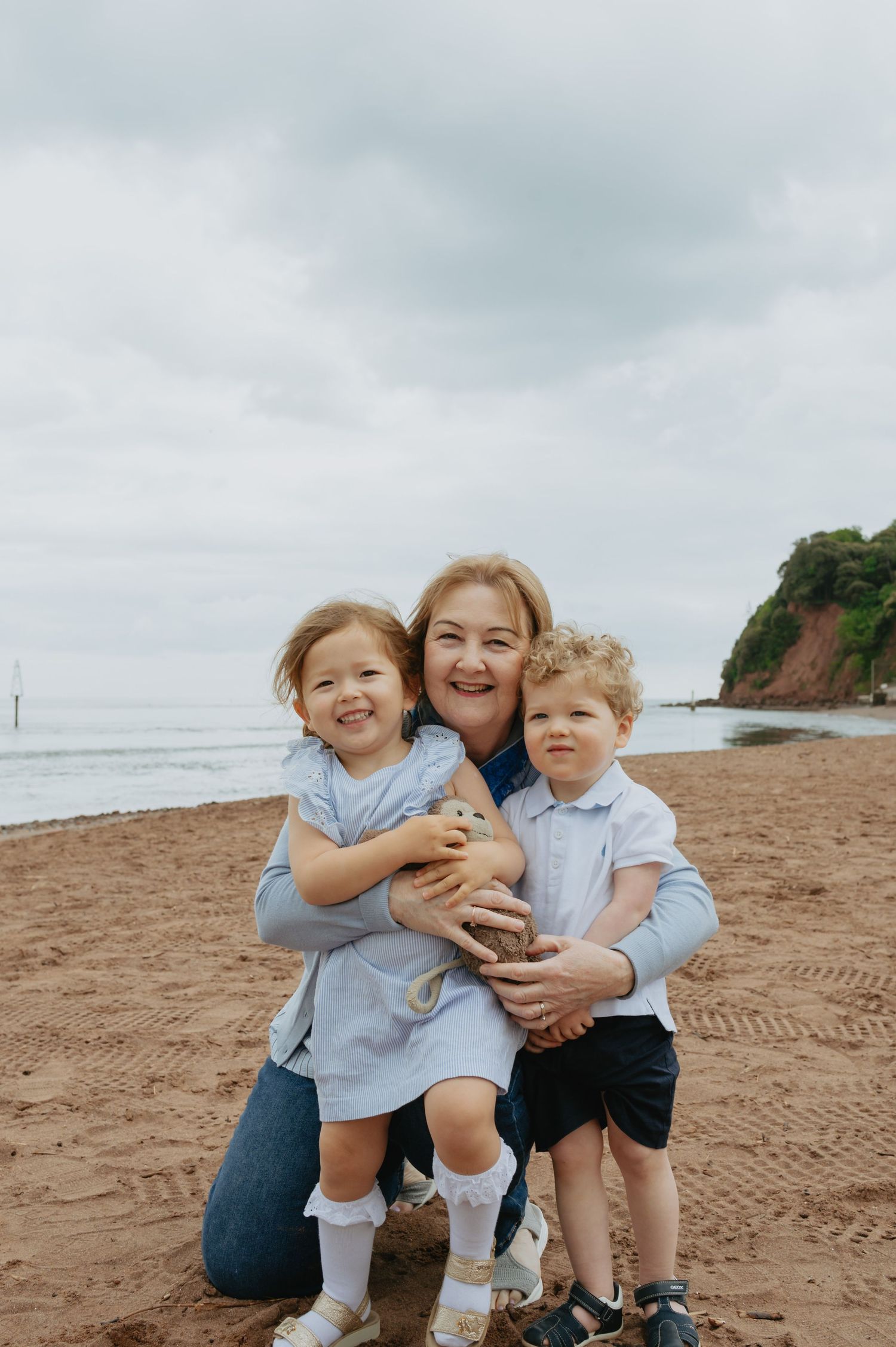 A grandmother hugs two young children on a sandy beach with a rocky headland and cloudy sky in the background.
