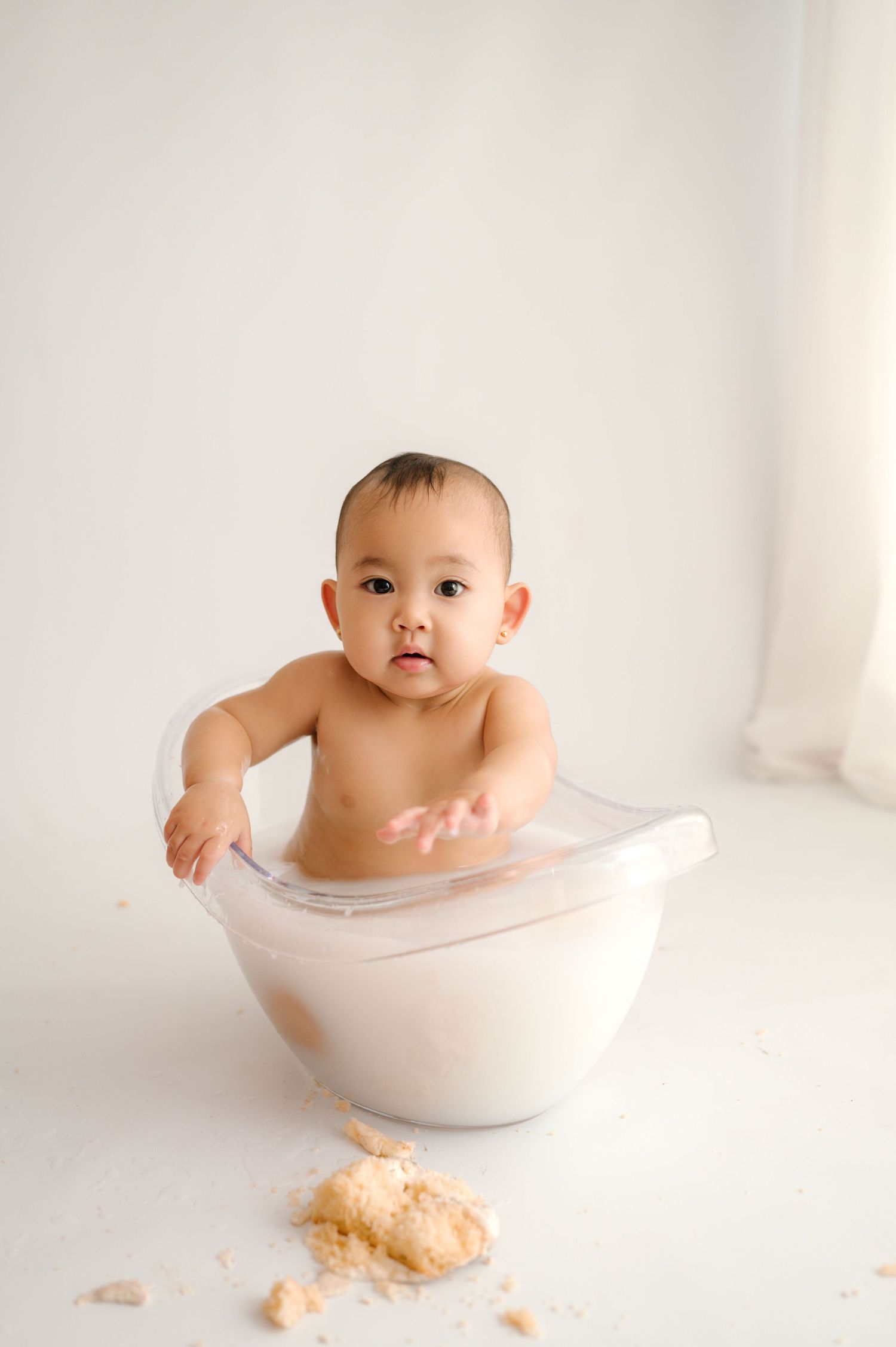 A cute baby sitting in a clear bowl with milk and cake crumbs on a white background.