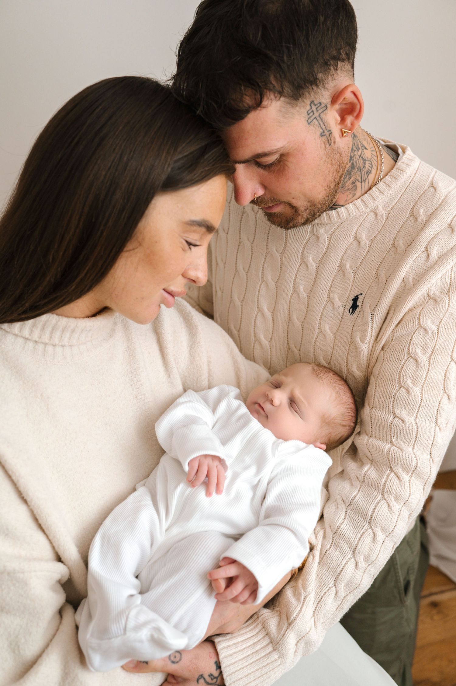 A young couple in matching cream cable-knit sweaters tenderly gazing at their sleeping newborn baby dressed in white.