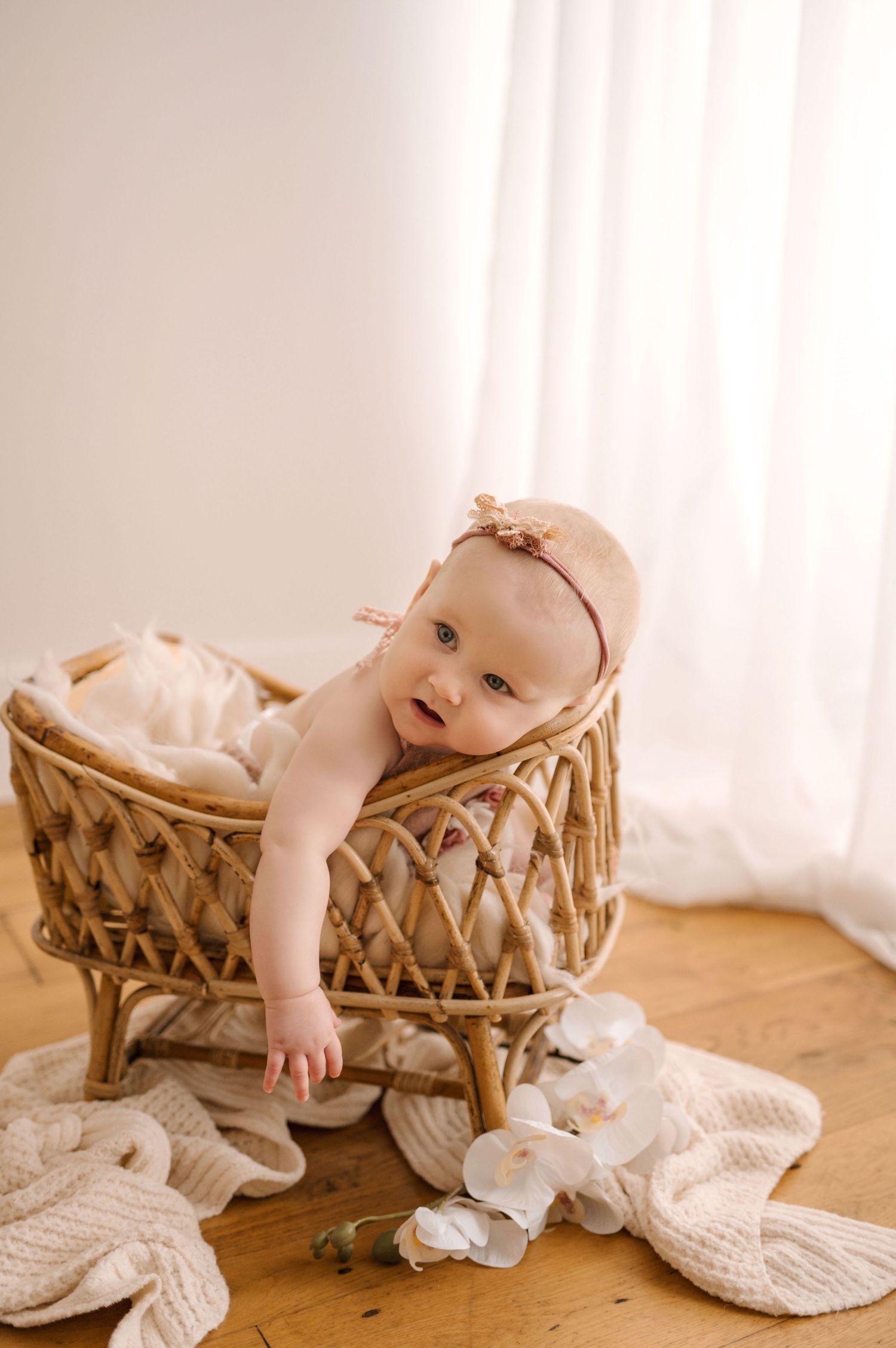 Baby in white outfit reaching over edge of woven basket on cream blanket during indoor portrait session.