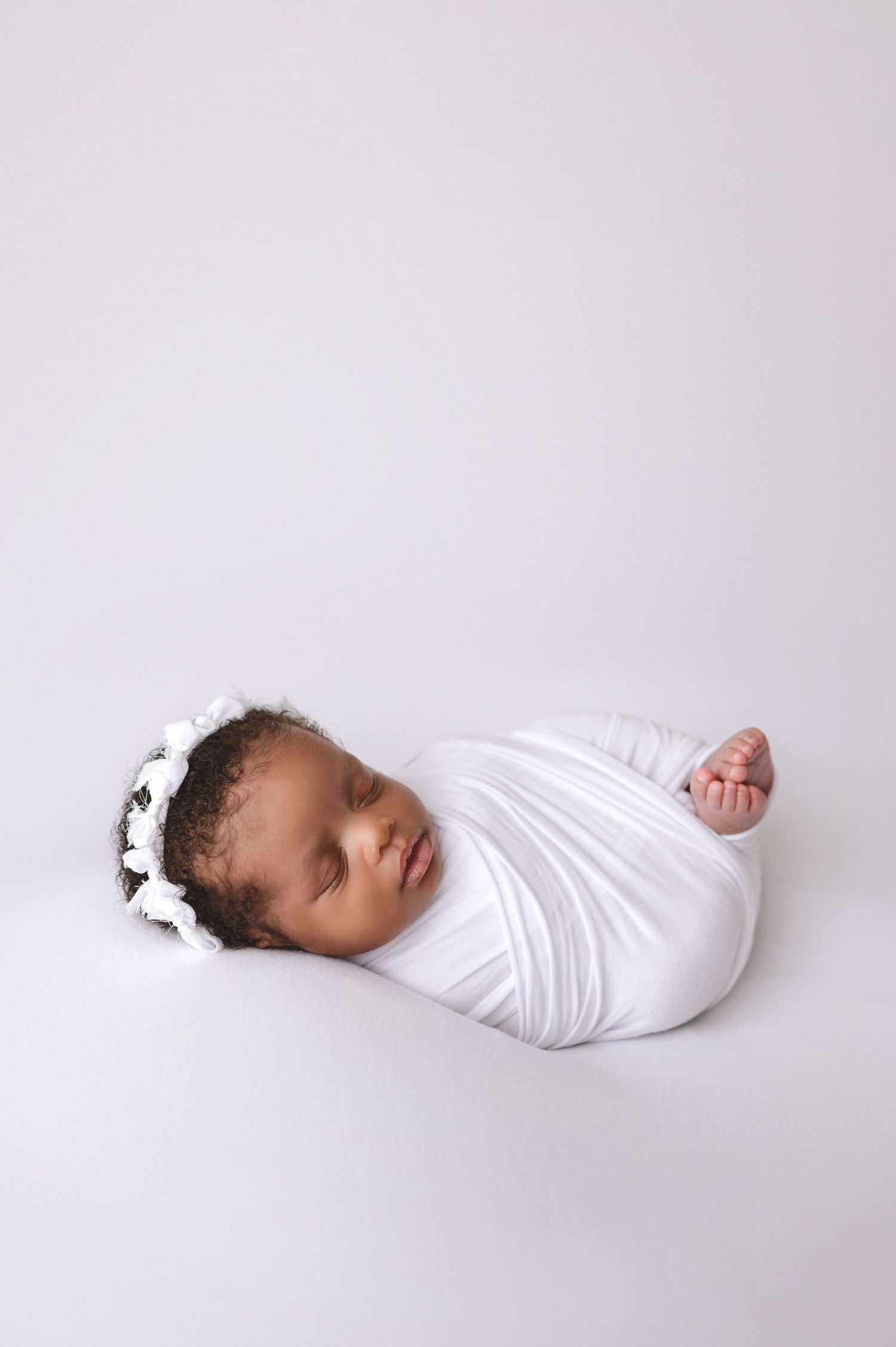 Peaceful sleeping newborn swaddled in white fabric with small floral crown against light background.