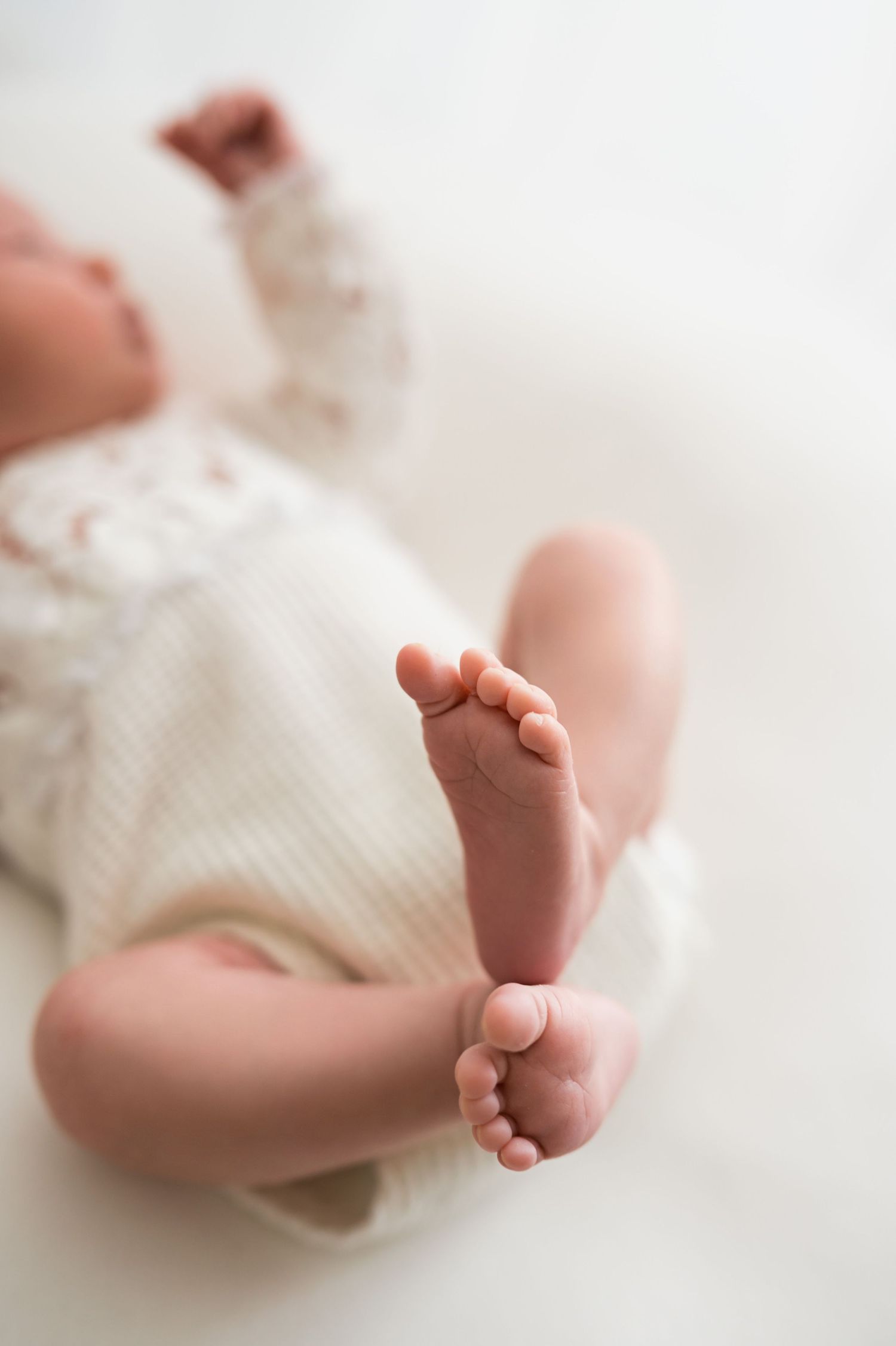 Close-up of tiny newborn baby feet in a white onesie lying on a soft white blanket.